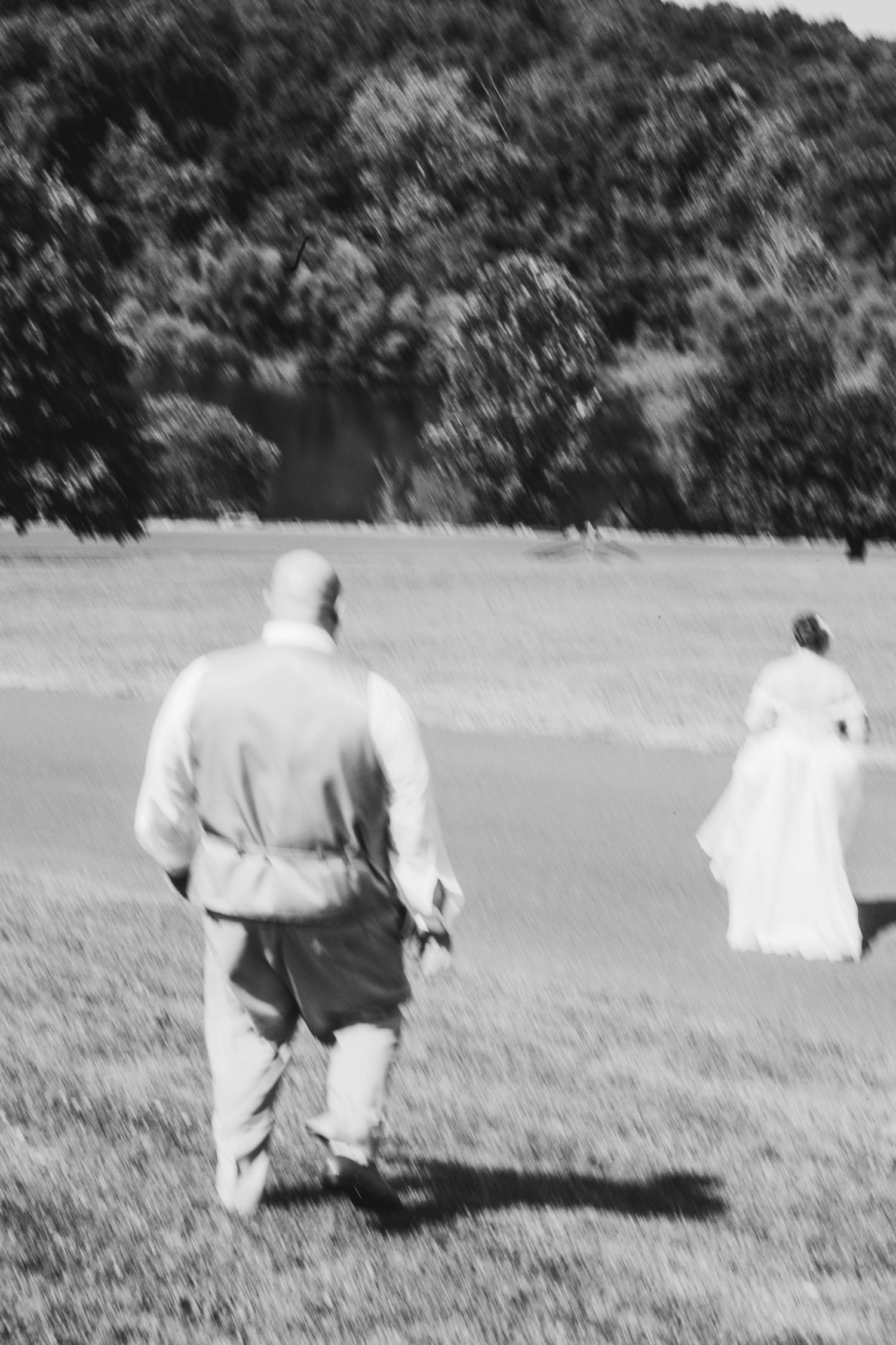 Black and white photo of bride and groom walking across green field at outdoor wedding.