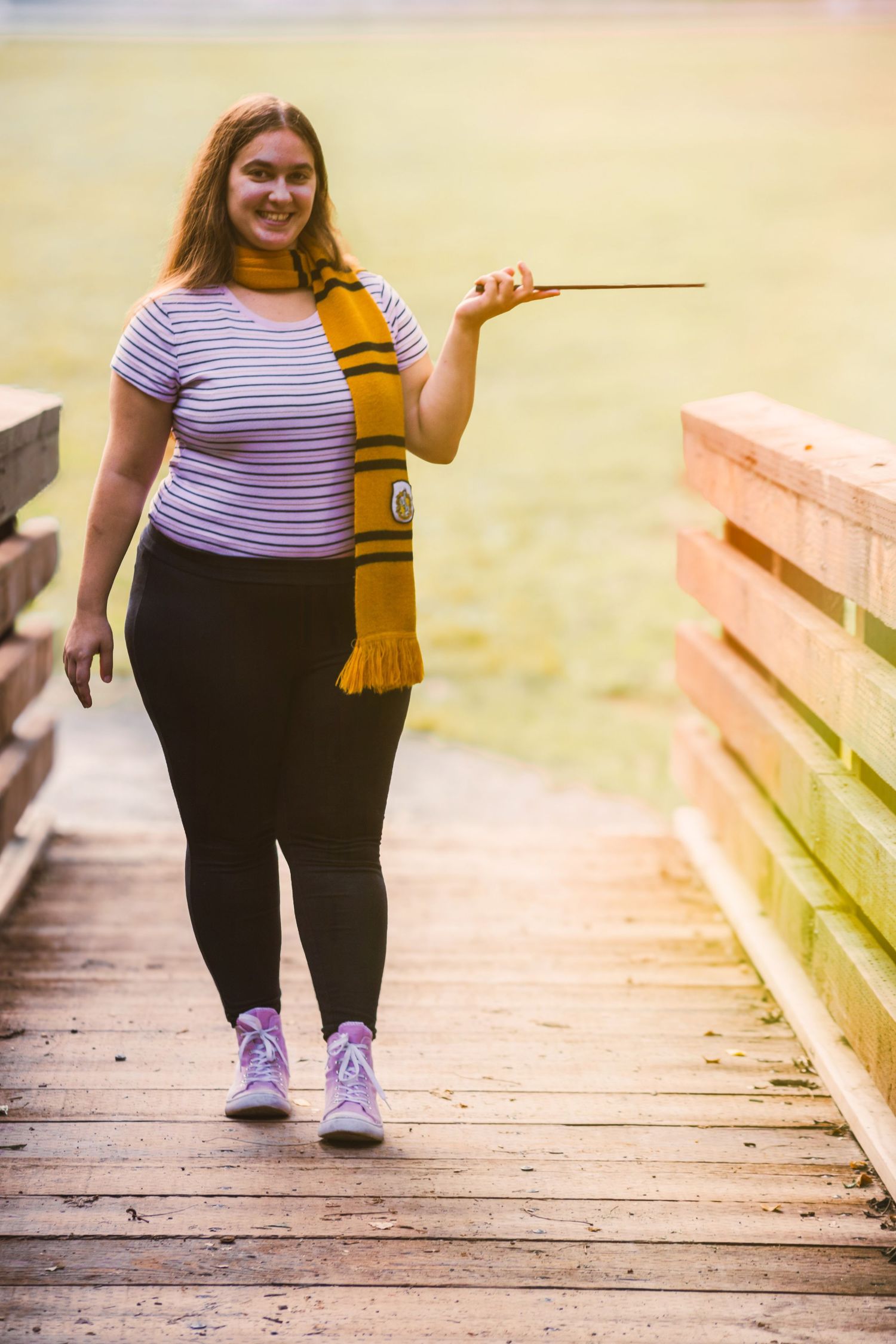 Young woman in Hufflepuff scarf holding a magic wand on a wooden bridge, cosplaying as a Harry Potter character.