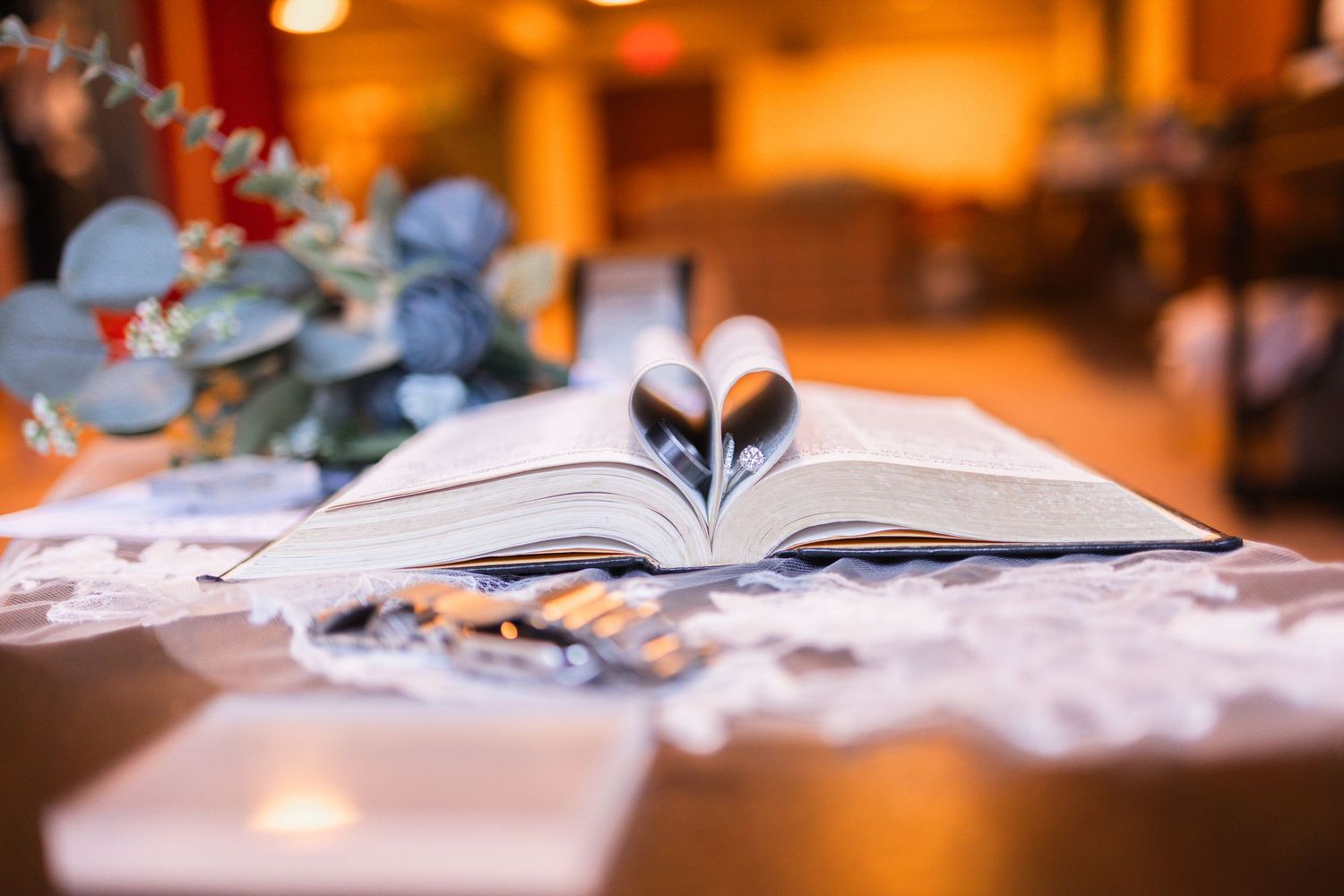 Open book with pages folded into a heart shape on a decorated table with floral arrangement in soft warm light.