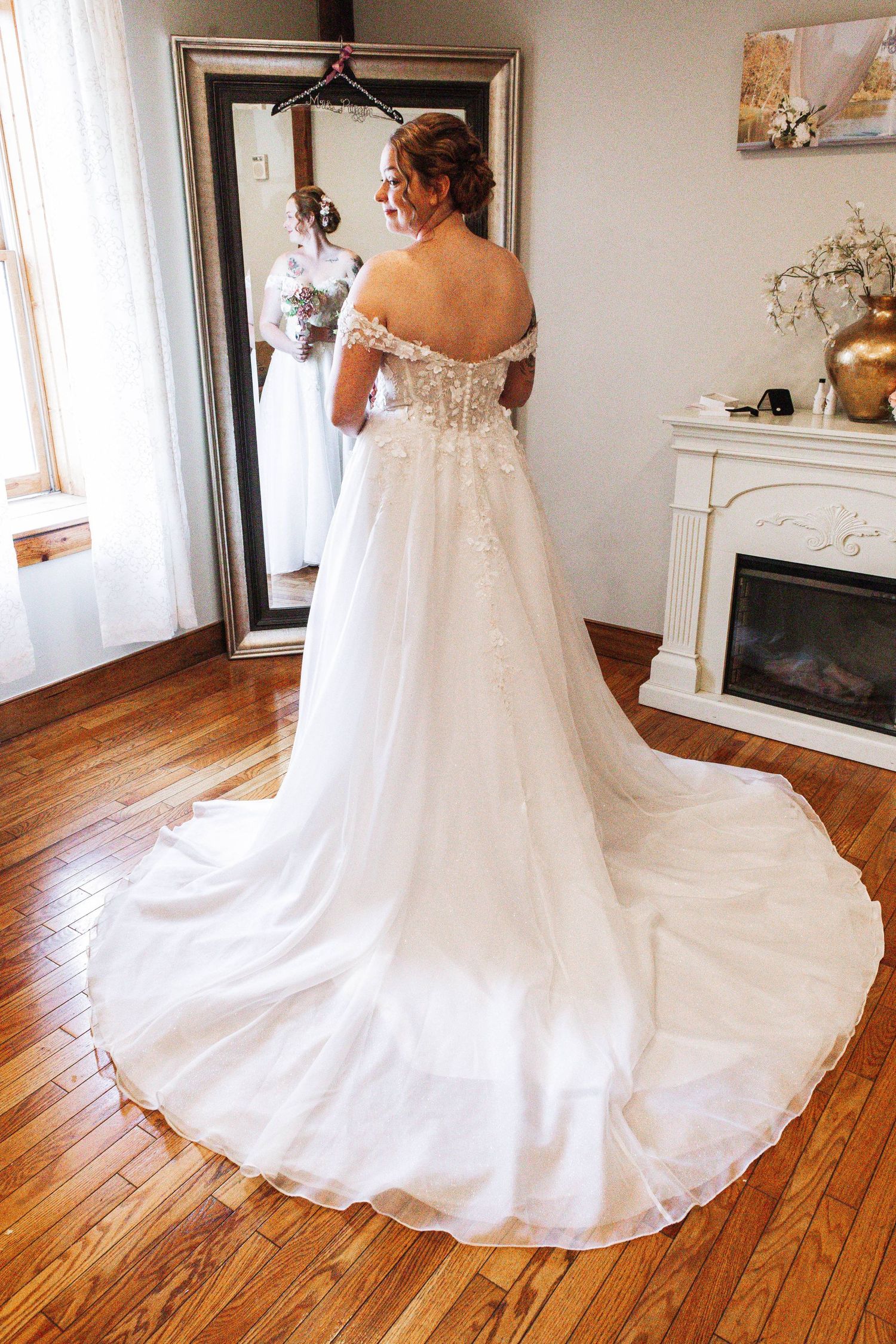 Bride in an off-shoulder lace wedding gown with a long train stands before a mirror.