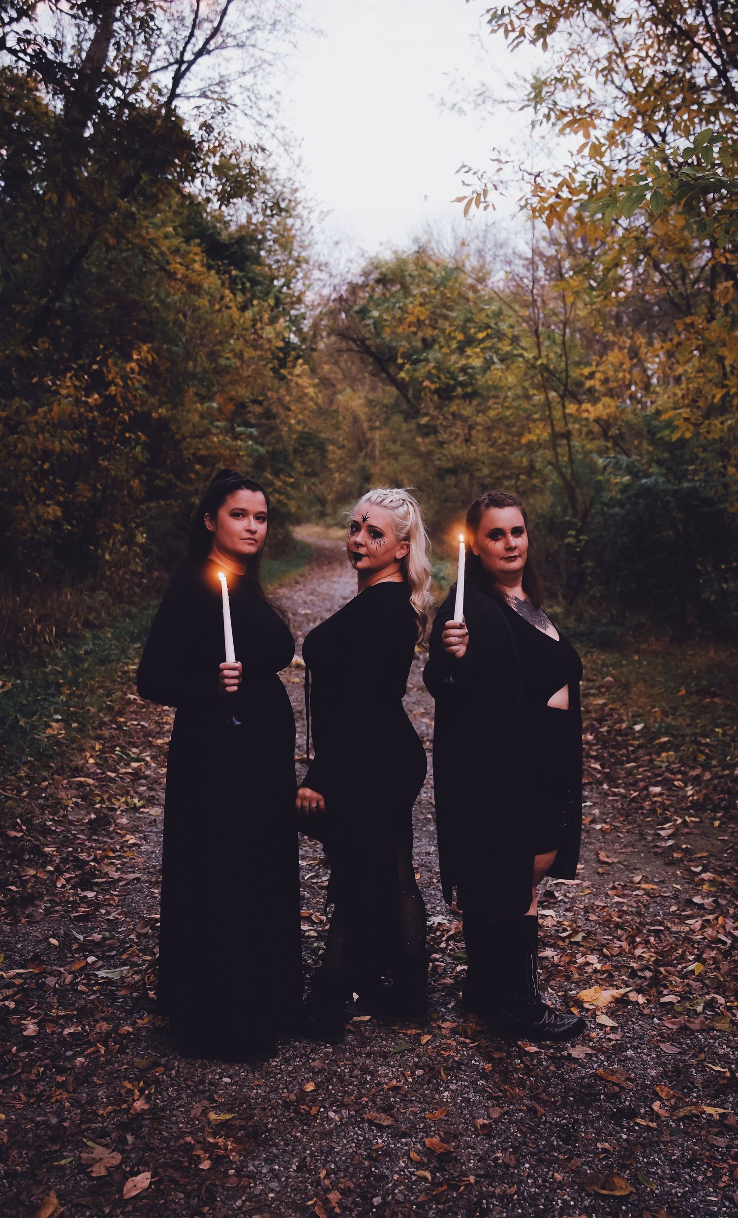Three women in black robes holding lit candles on an autumn forest path at dusk.