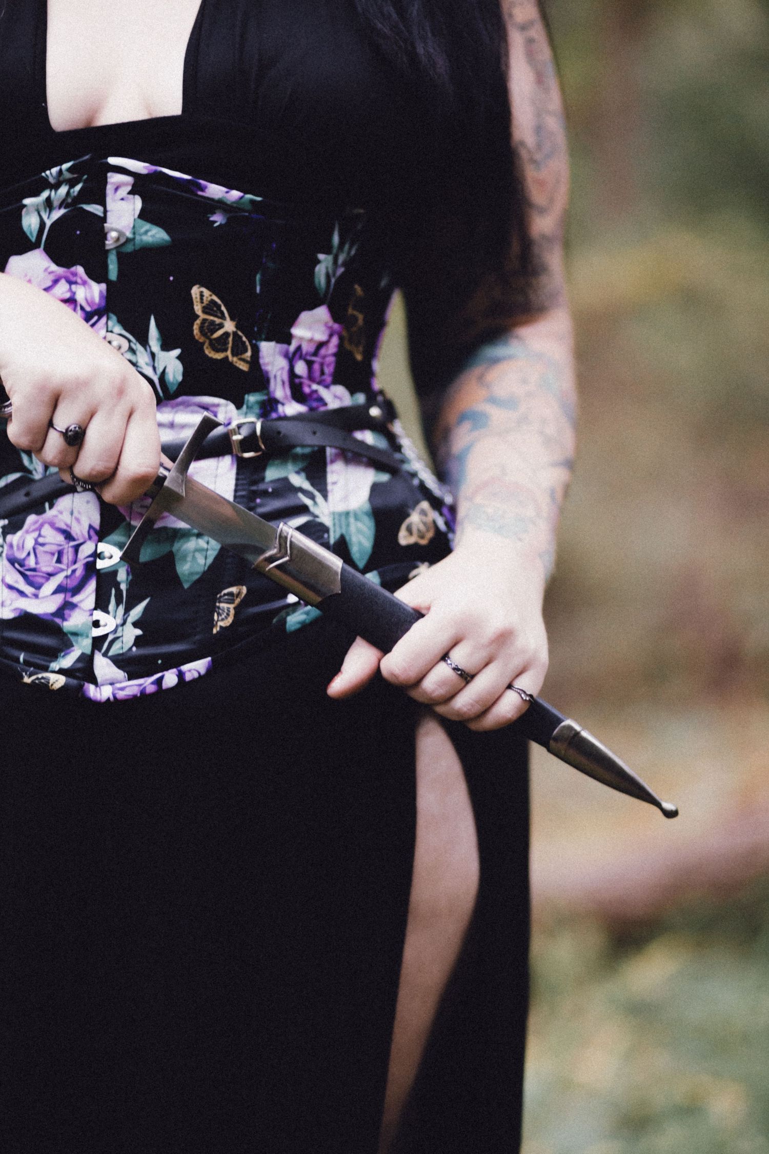 Tattooed woman in black floral dress holding a decorative dagger knife outdoors.