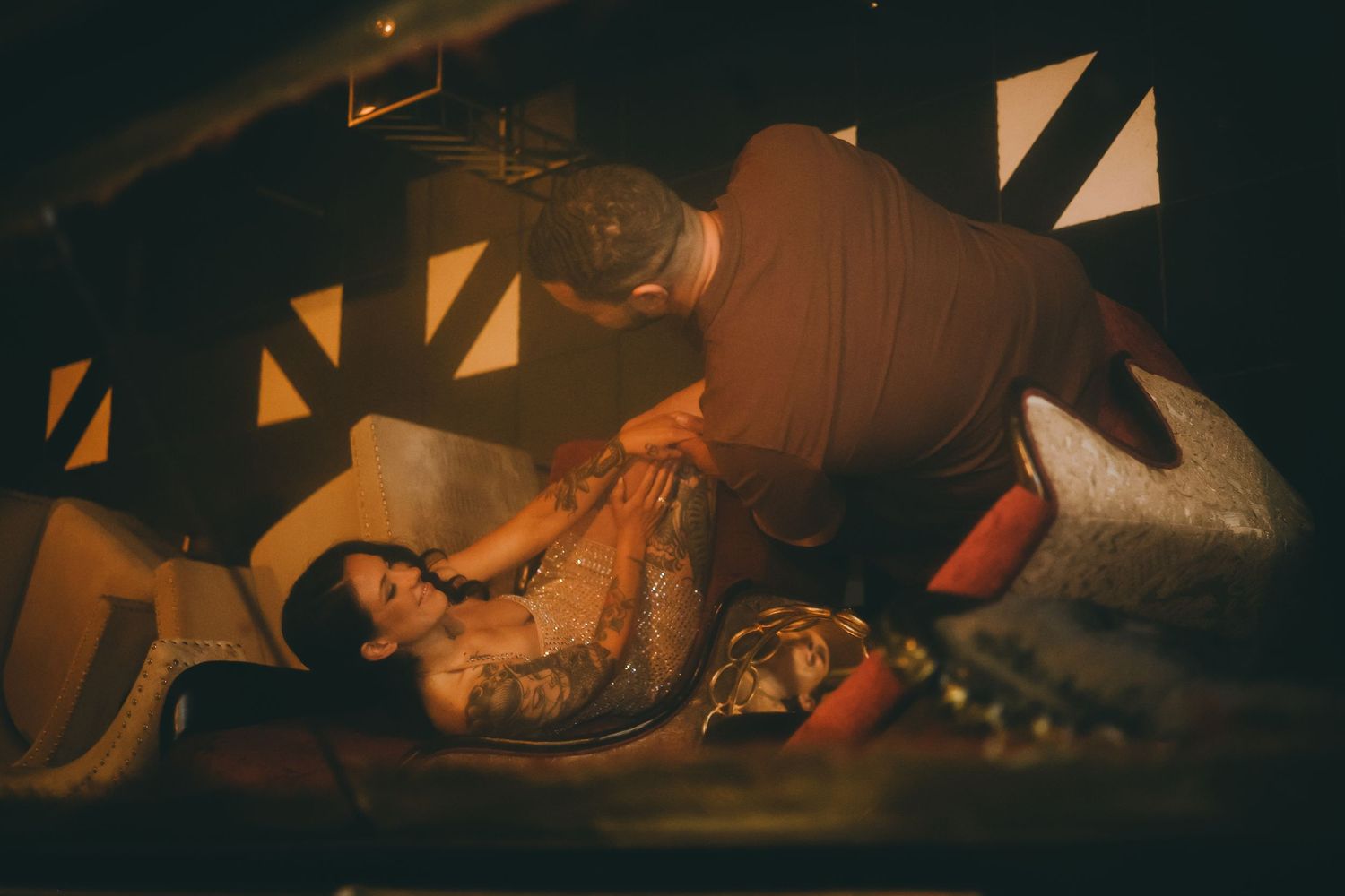 A tattooed woman reclines as a man leans over her in a dimly lit, dramatically decorated room.