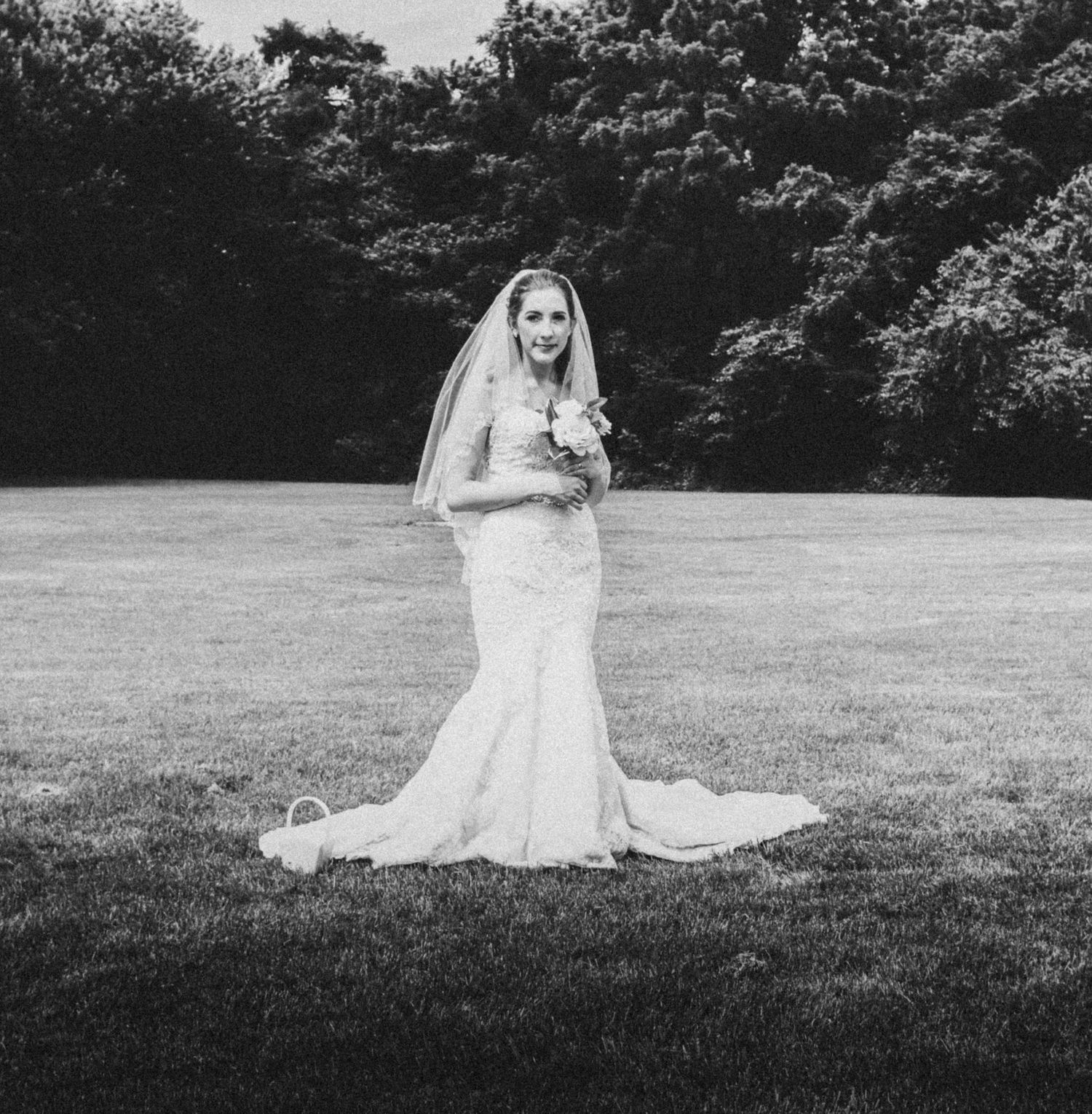 Black and white photo of a bride in a lace mermaid gown and veil holding a bouquet on a green lawn.