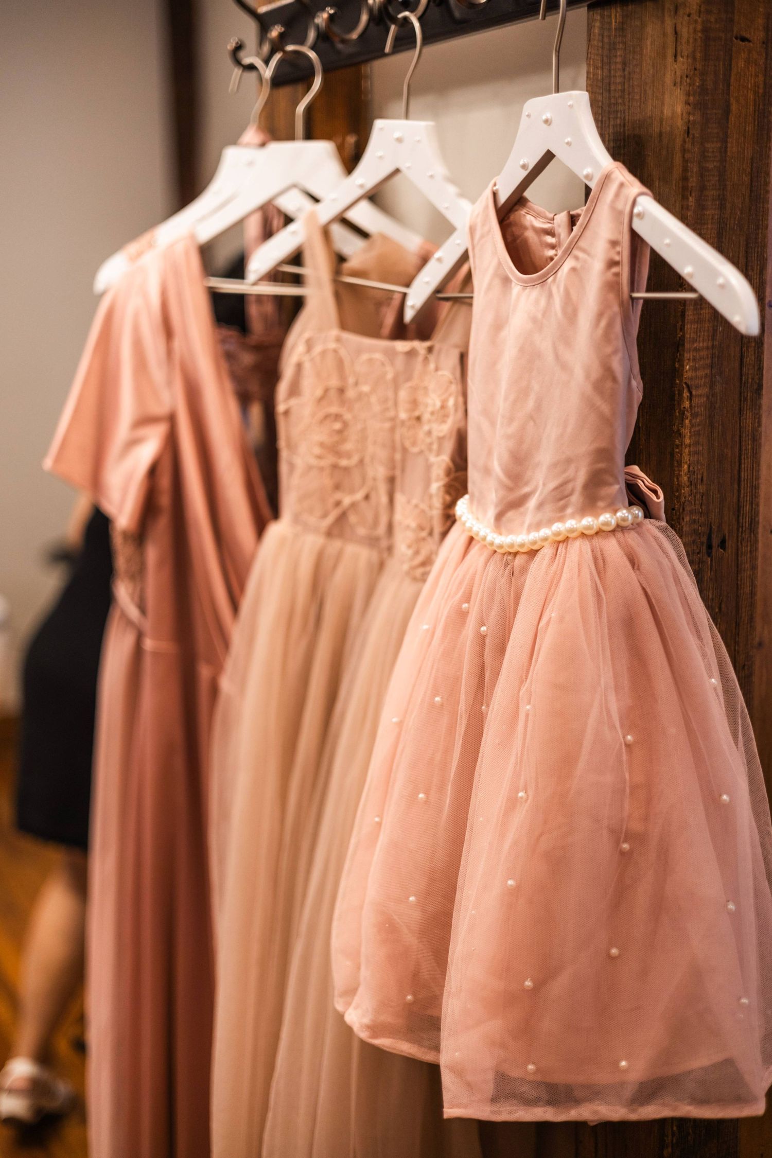 Three pink tulle flower girl dresses hanging on white hangers on a wooden rack.