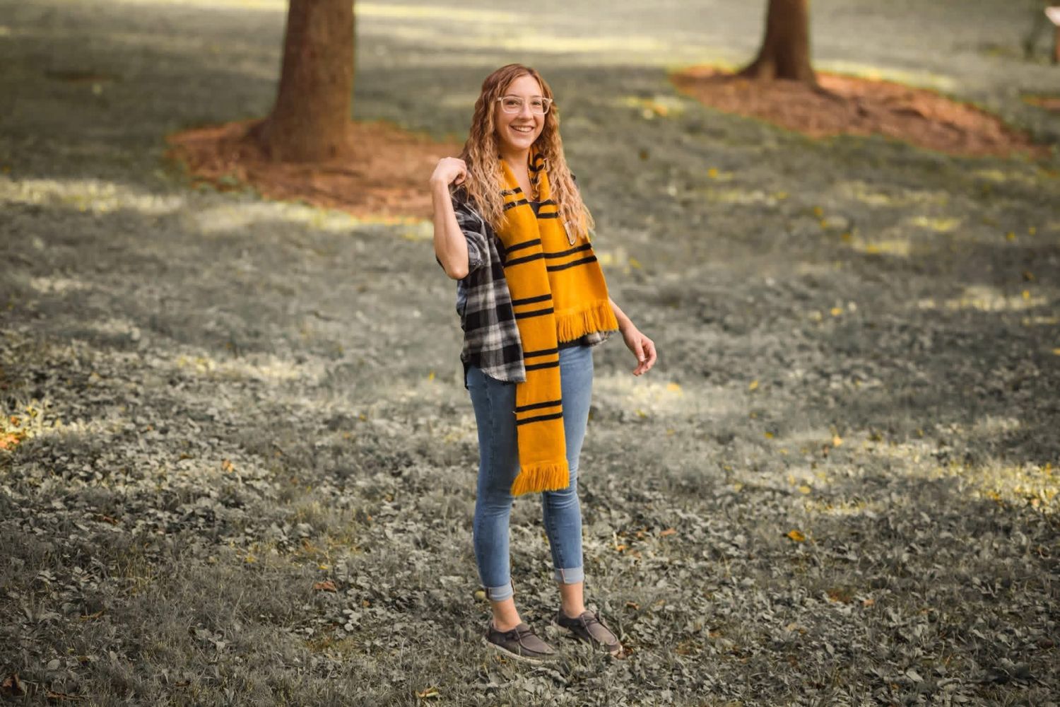 Pregnant woman in mustard yellow top and plaid vest smiling outdoors in a sunlit park setting.