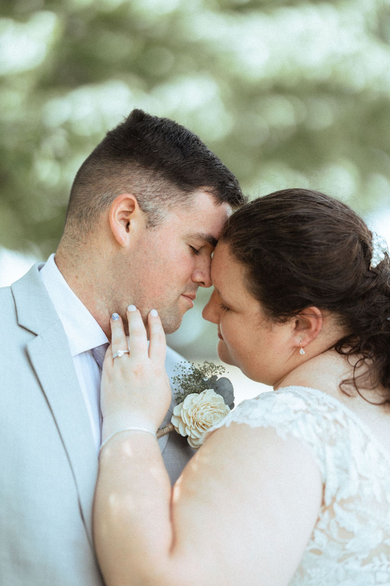 A bride and groom share an intimate forehead touch moment, dressed in wedding attire with a boutonniere and lace dress.