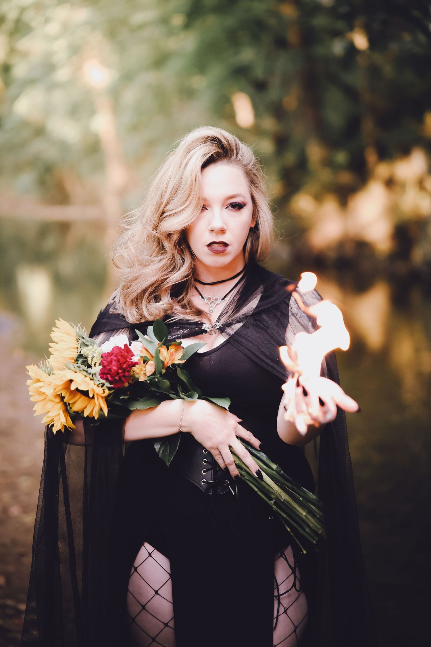 Blonde woman in black gothic costume holding sunflowers and a flame in an outdoor autumn forest setting.