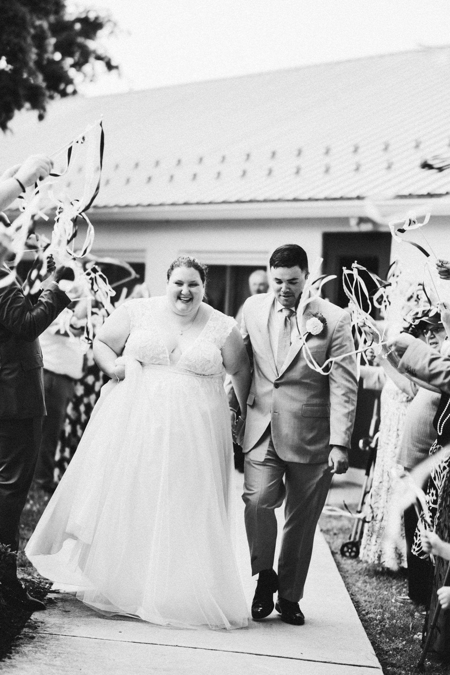 Black and white photo of a smiling bride and groom walking through a ribbon wand send-off after their wedding ceremony.