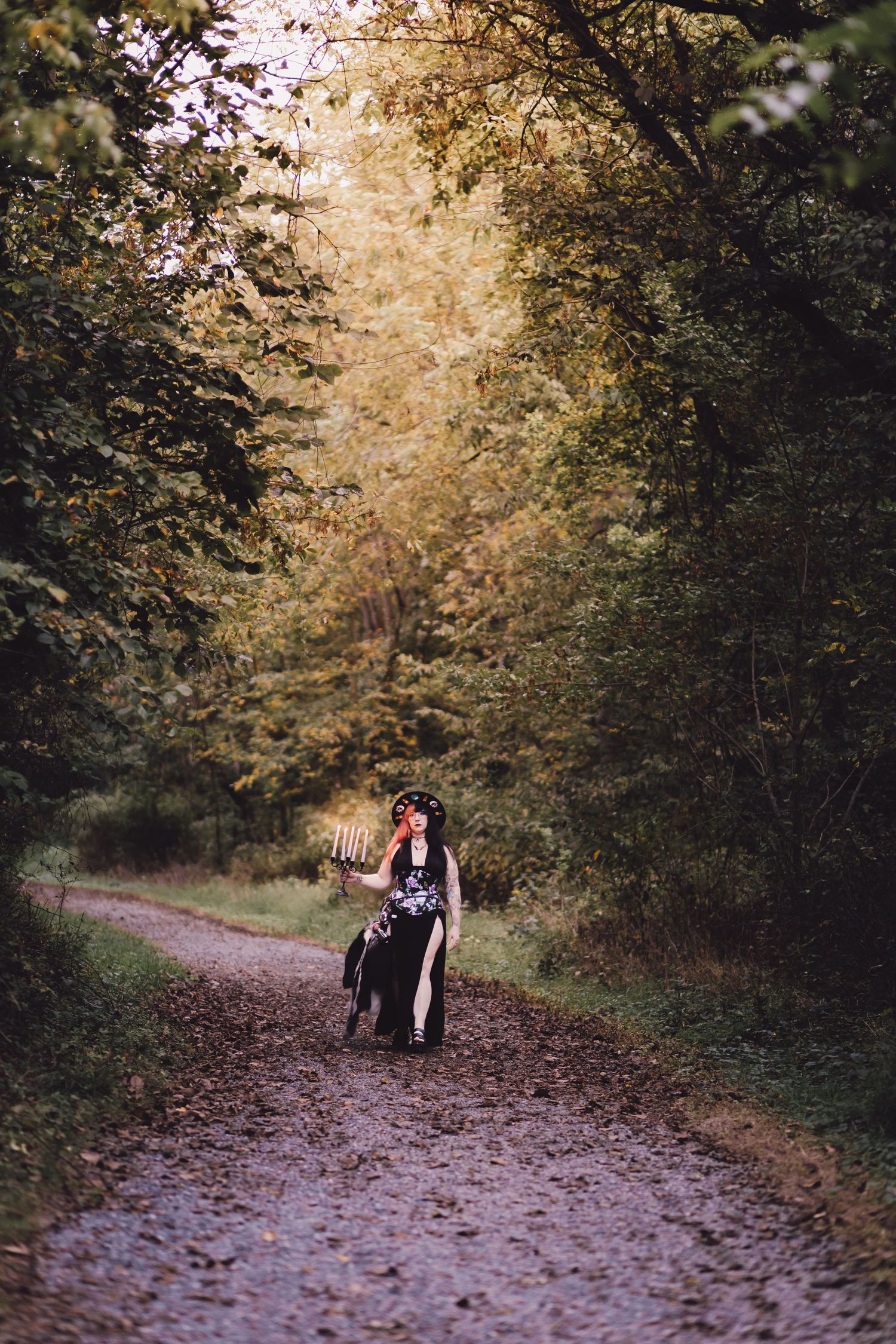 Woman in black outfit walking a dog down a leaf-covered forest path in autumn.