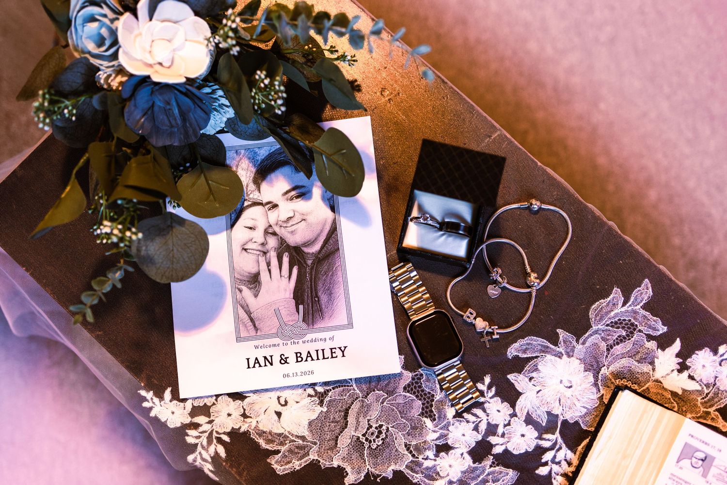 Ian and Bailey wedding flat lay with photo card, blue flowers, lace, rings, and bracelet on a table.