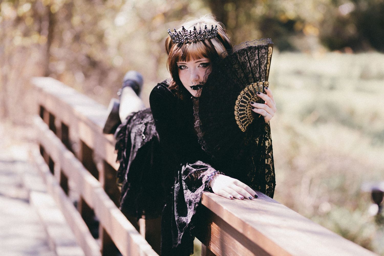 Woman in gothic black lace dress and crown holds decorative fan on wooden bridge in autumn setting.