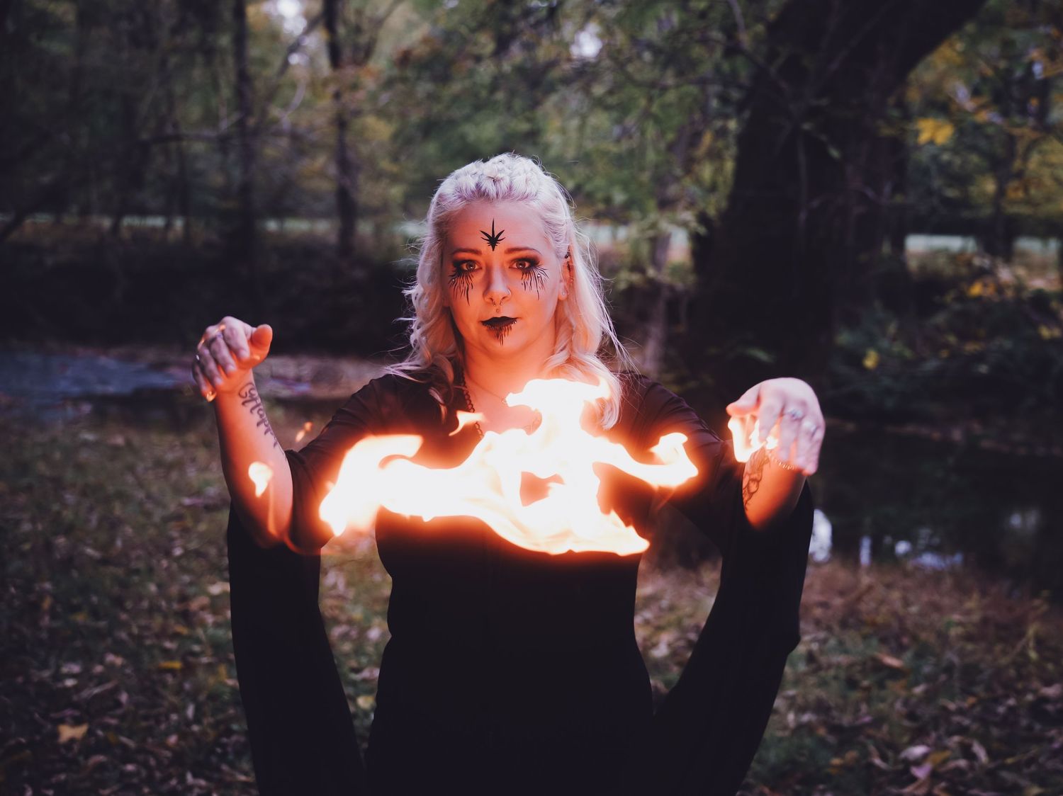 Woman in black dress holding fire with cross face paint in dark forest setting.