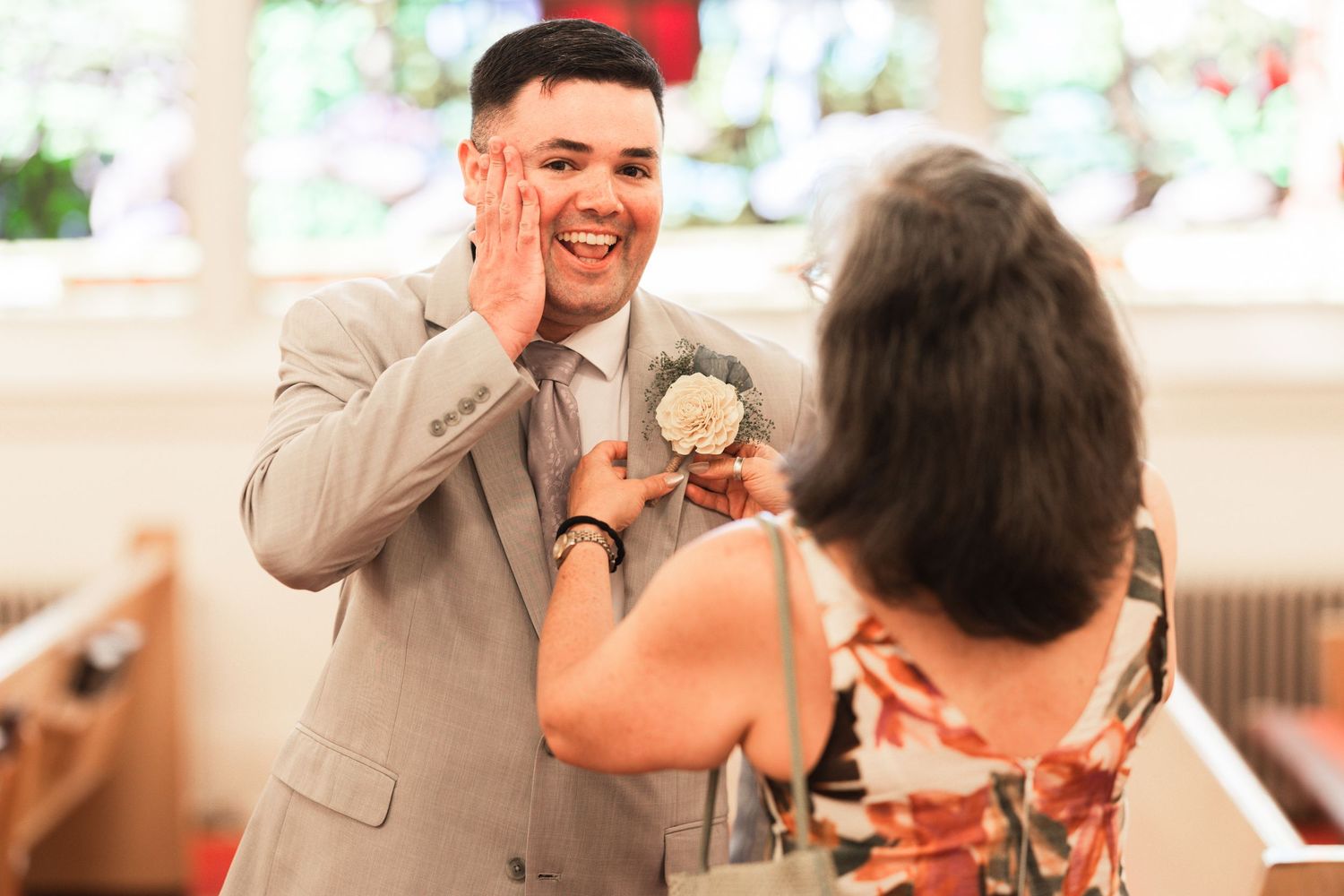 Groom in gray suit laughing as woman pins boutonniere at wedding ceremony.