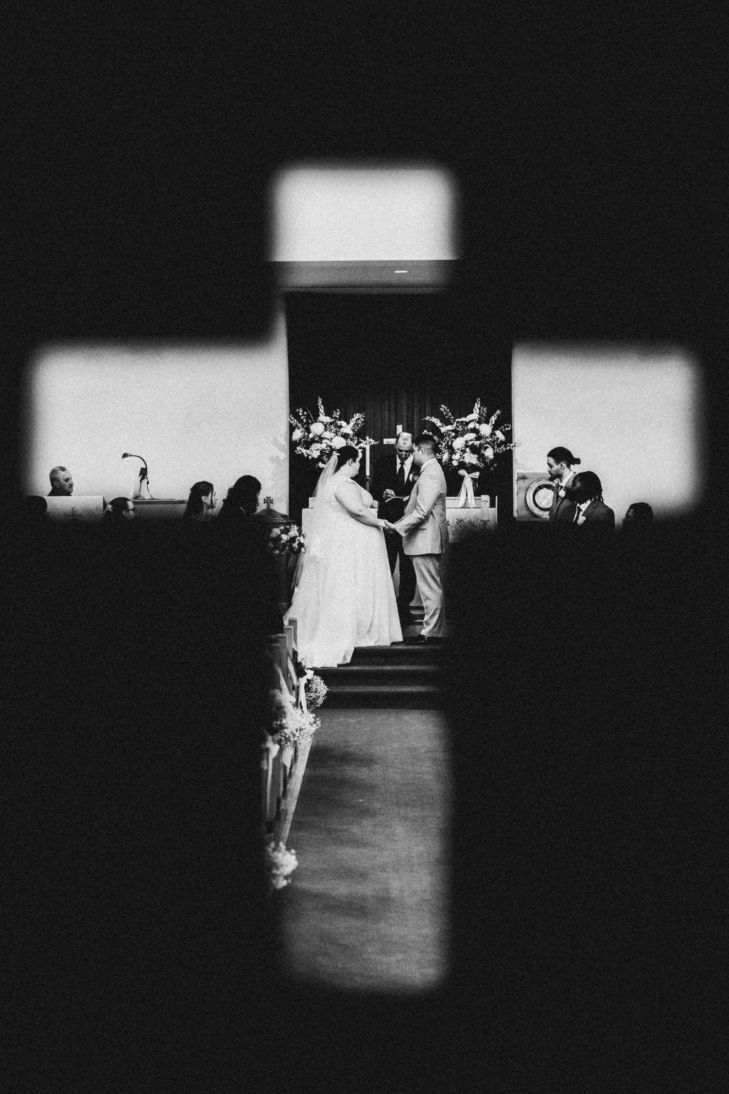 Black and white photo of a wedding ceremony framed through a cross-shaped window in a church.