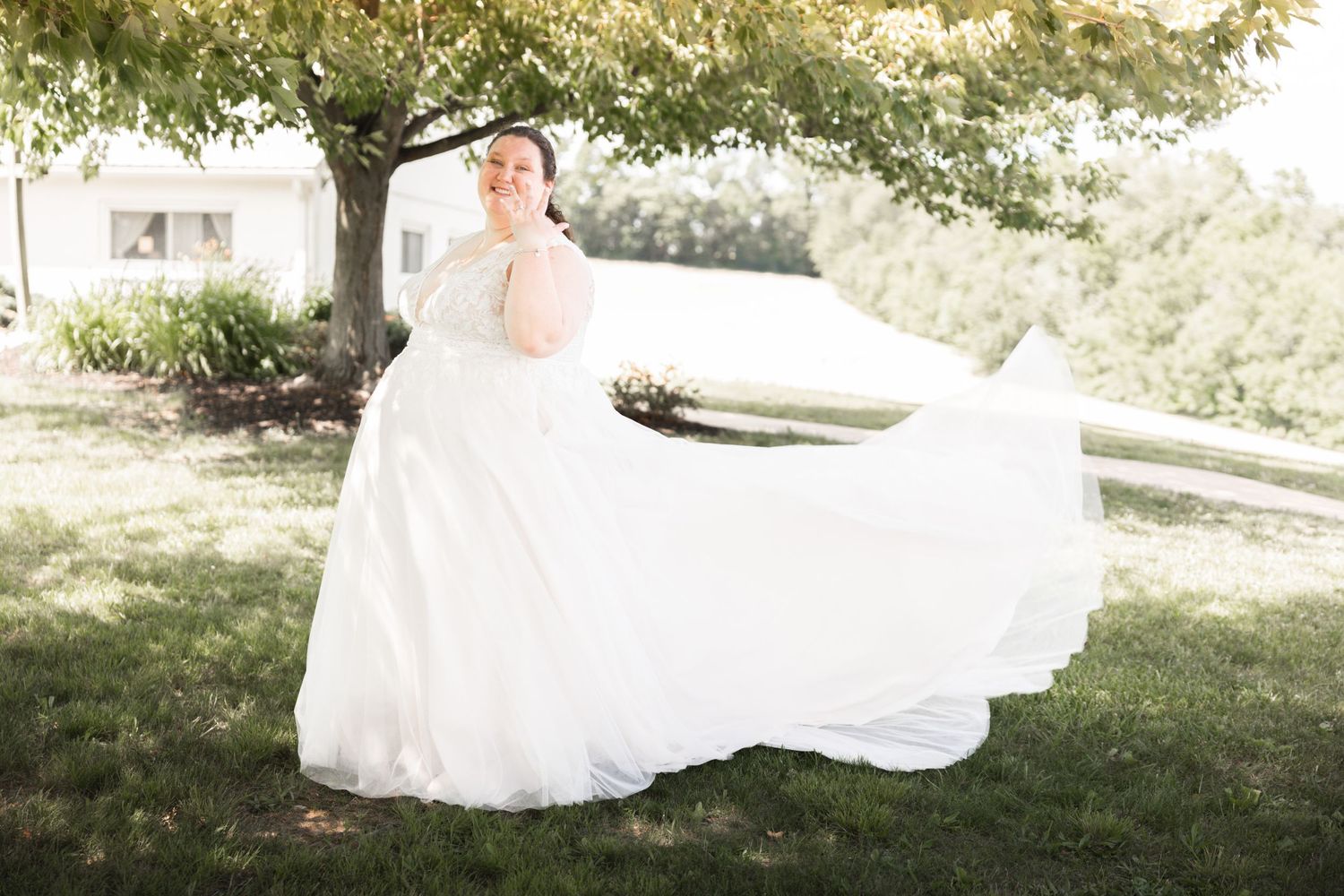Bride in flowing white wedding gown twirling outdoors under a tree on a sunny day.