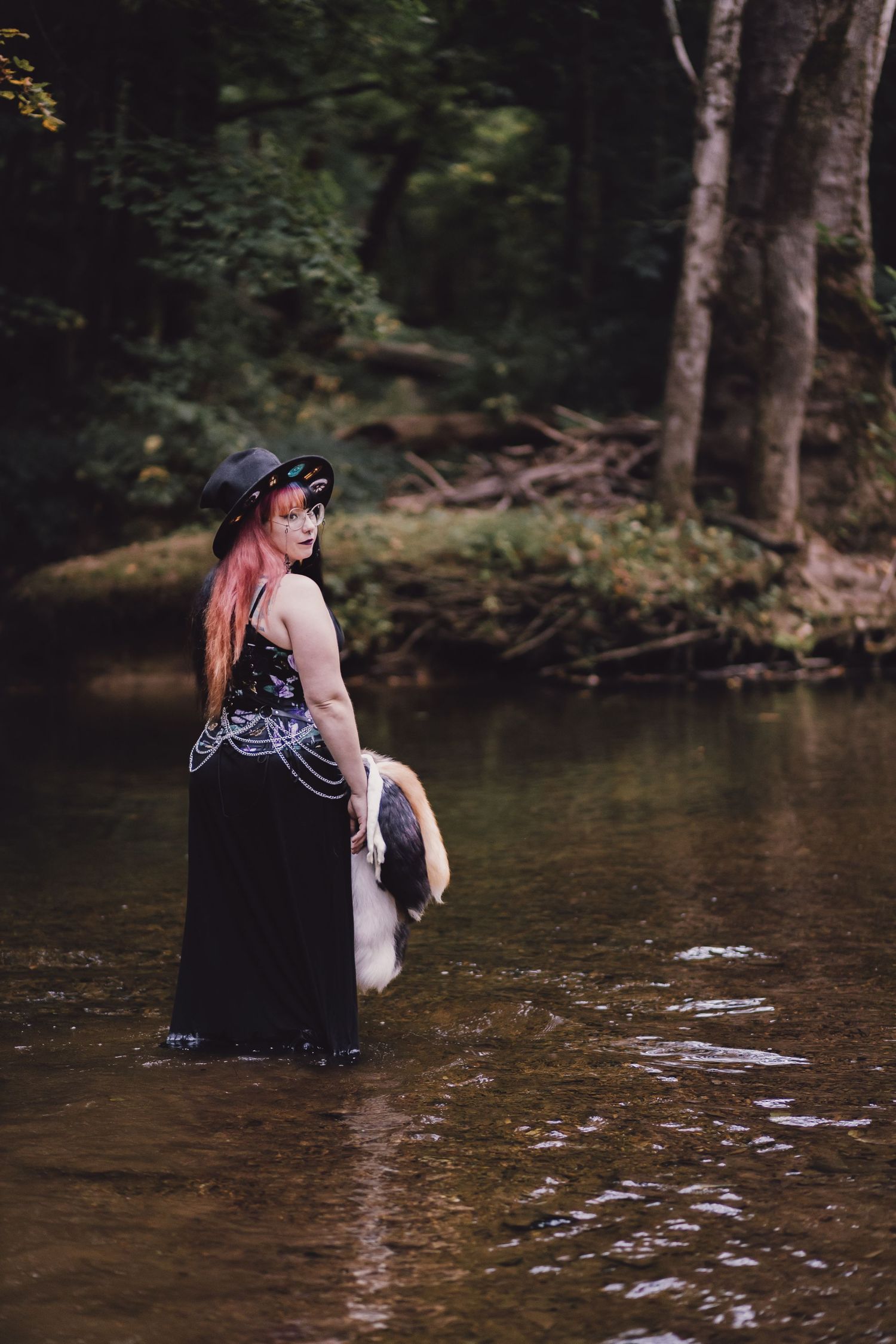 Woman in black gothic dress and hat standing in a shallow forest creek holding white fabric.