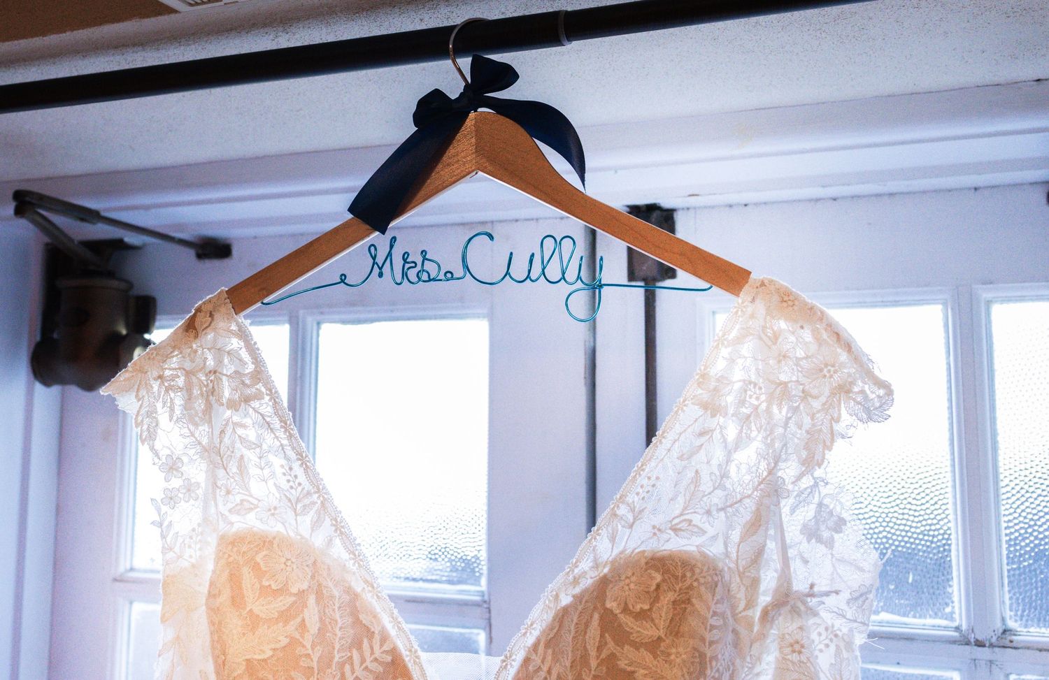 Wedding dress on a personalized Mrs. Cully wire hanger displayed near a bright window.