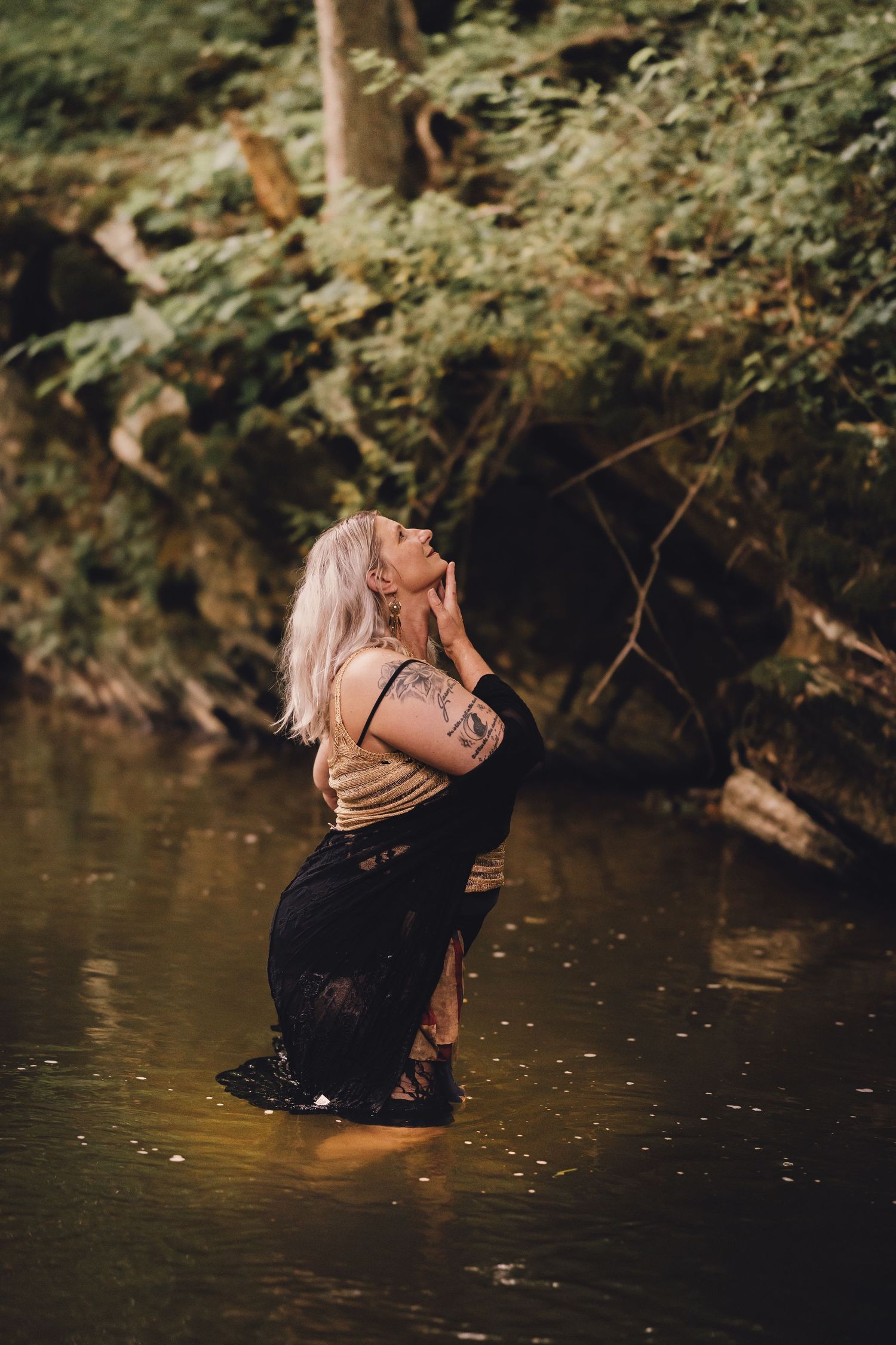 A tattooed woman with pink hair stands in a forest stream wearing a black skirt, head tilted back in a serene pose.
