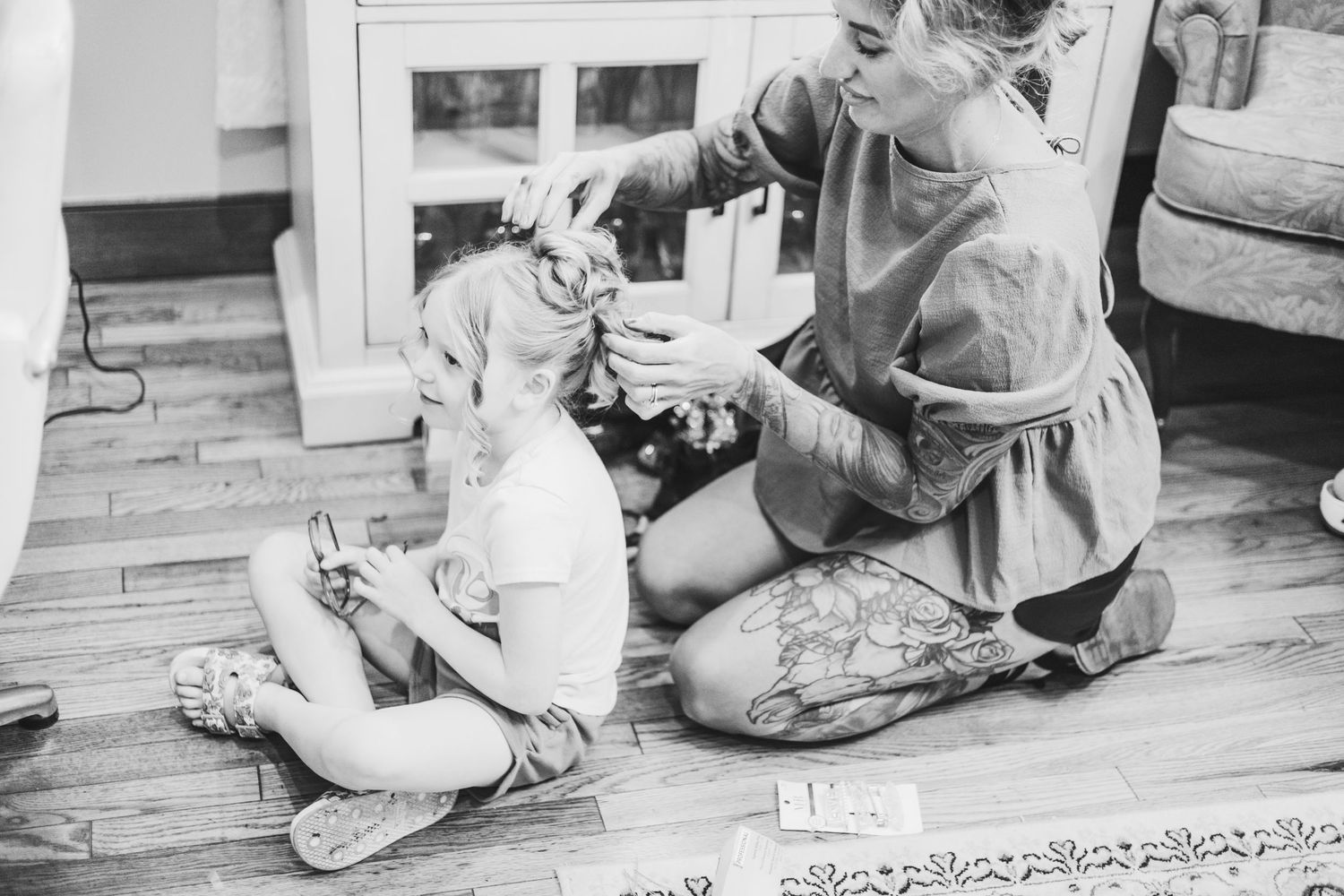 Black and white photo of a mother styling her young daughter's hair while sitting on a hardwood floor.
