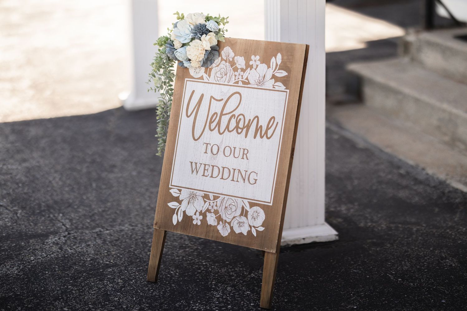 Wooden A-frame welcome to our wedding sign decorated with white floral arrangements on black pavement.