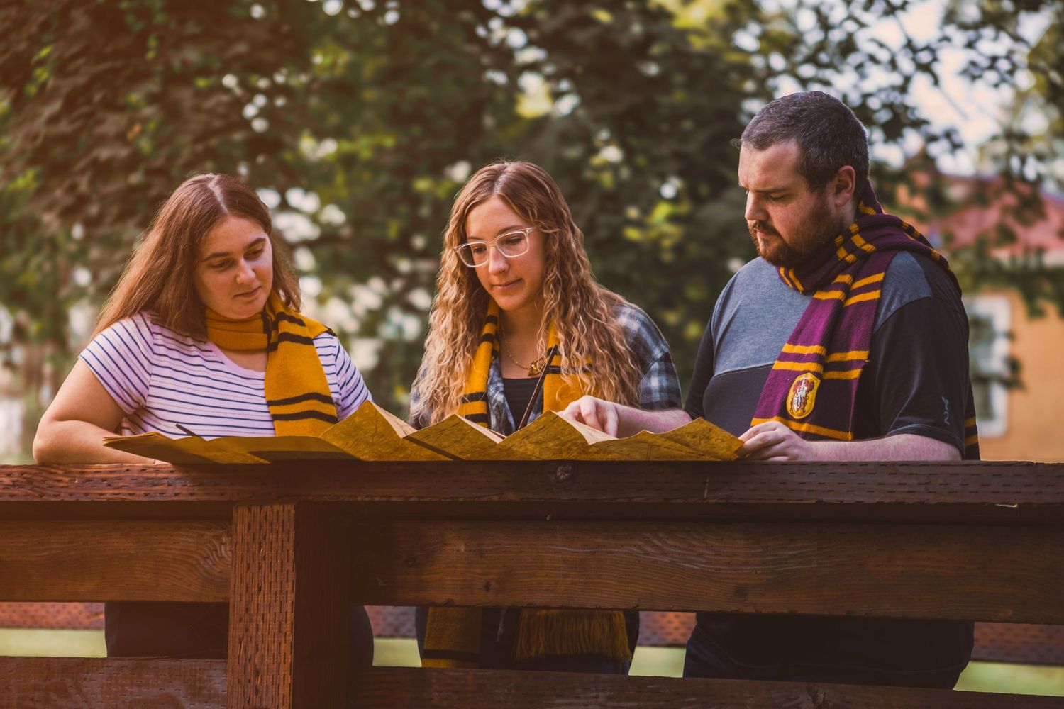 Three students in Hogwarts scarves study together at an outdoor wooden table surrounded by trees.