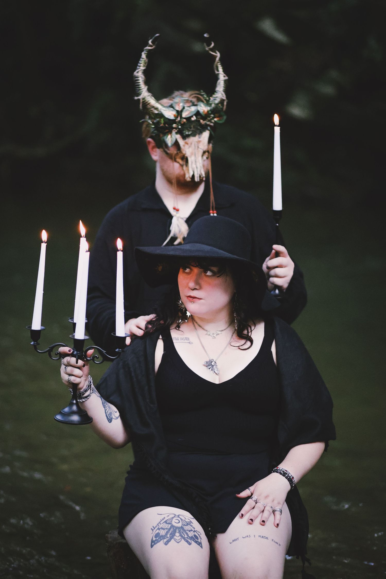 Gothic couple in black clothing holding candles with skull horned crown in dark setting.