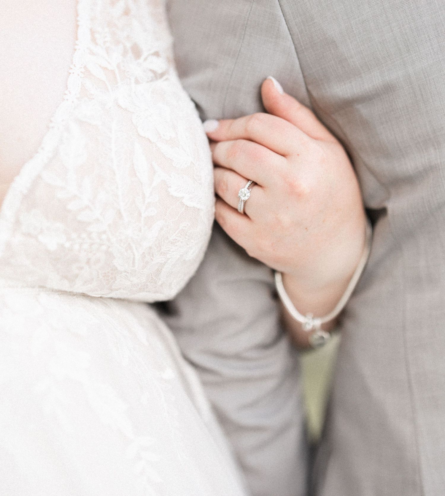Close-up of bride in lace wedding dress and groom in gray suit, showcasing elegant diamond engagement ring on bride's hand.