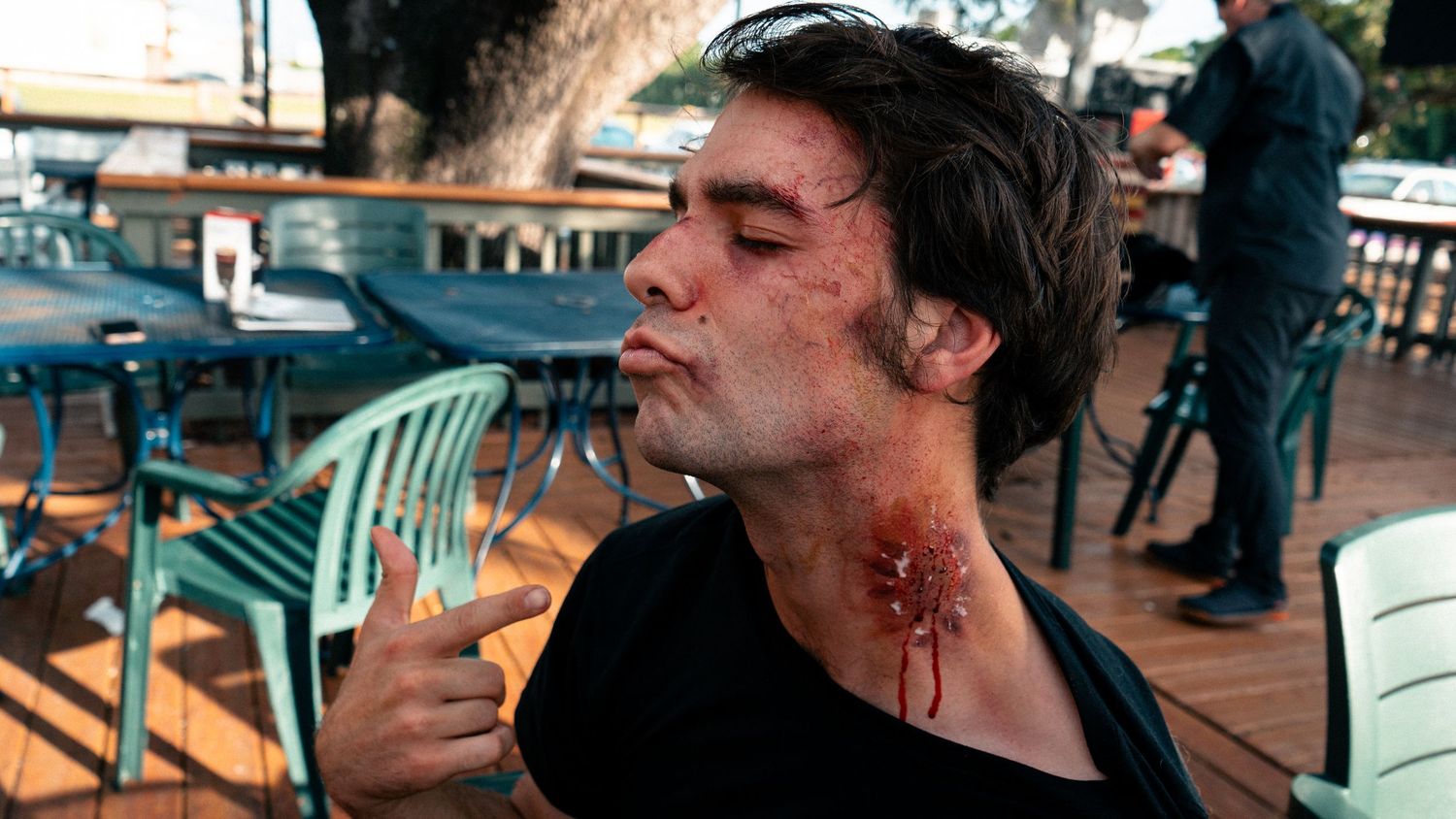 Young man with realistic bloody wound makeup on face and neck poses outdoors at a patio restaurant.