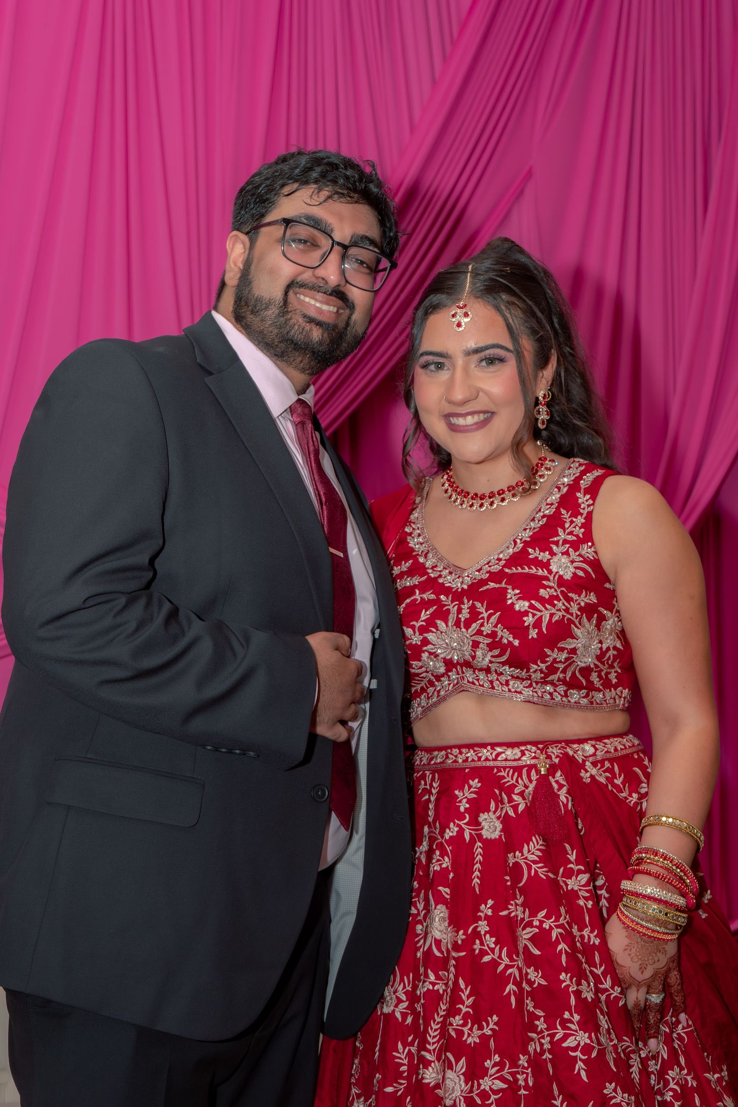 Couple posing at Indian wedding, man in dark suit with red tie, woman in red embroidered lehenga with jewelry, pink backdrop.