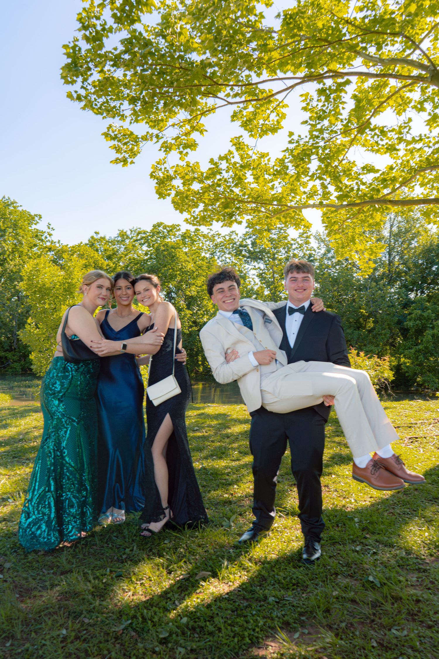 Group of prom-dressed teens pose humorously as one young man in a white suit is carried by another outdoors.