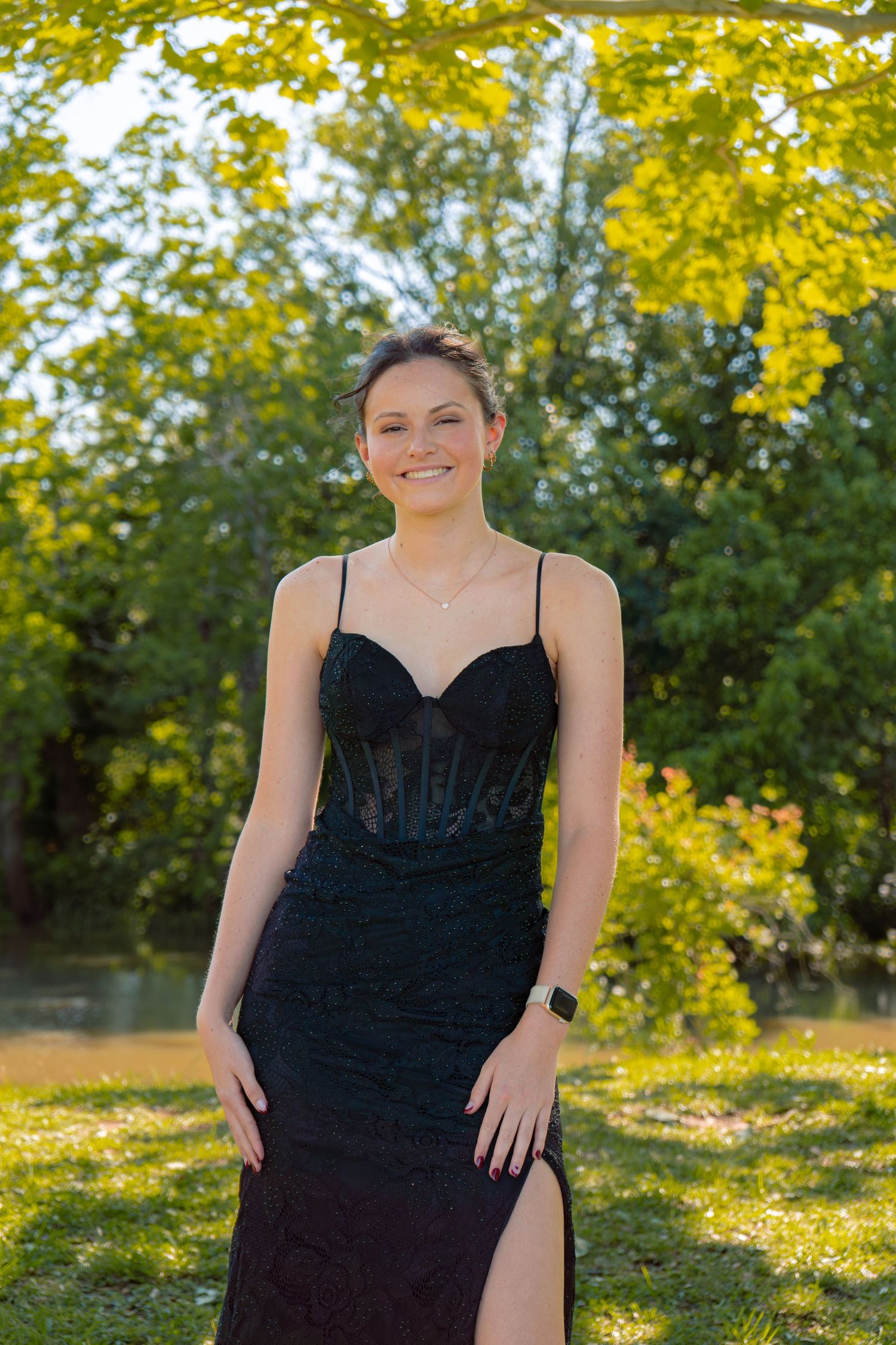 Young woman smiling in elegant black lace spaghetti strap gown with slit, posing outdoors in sunlit park.