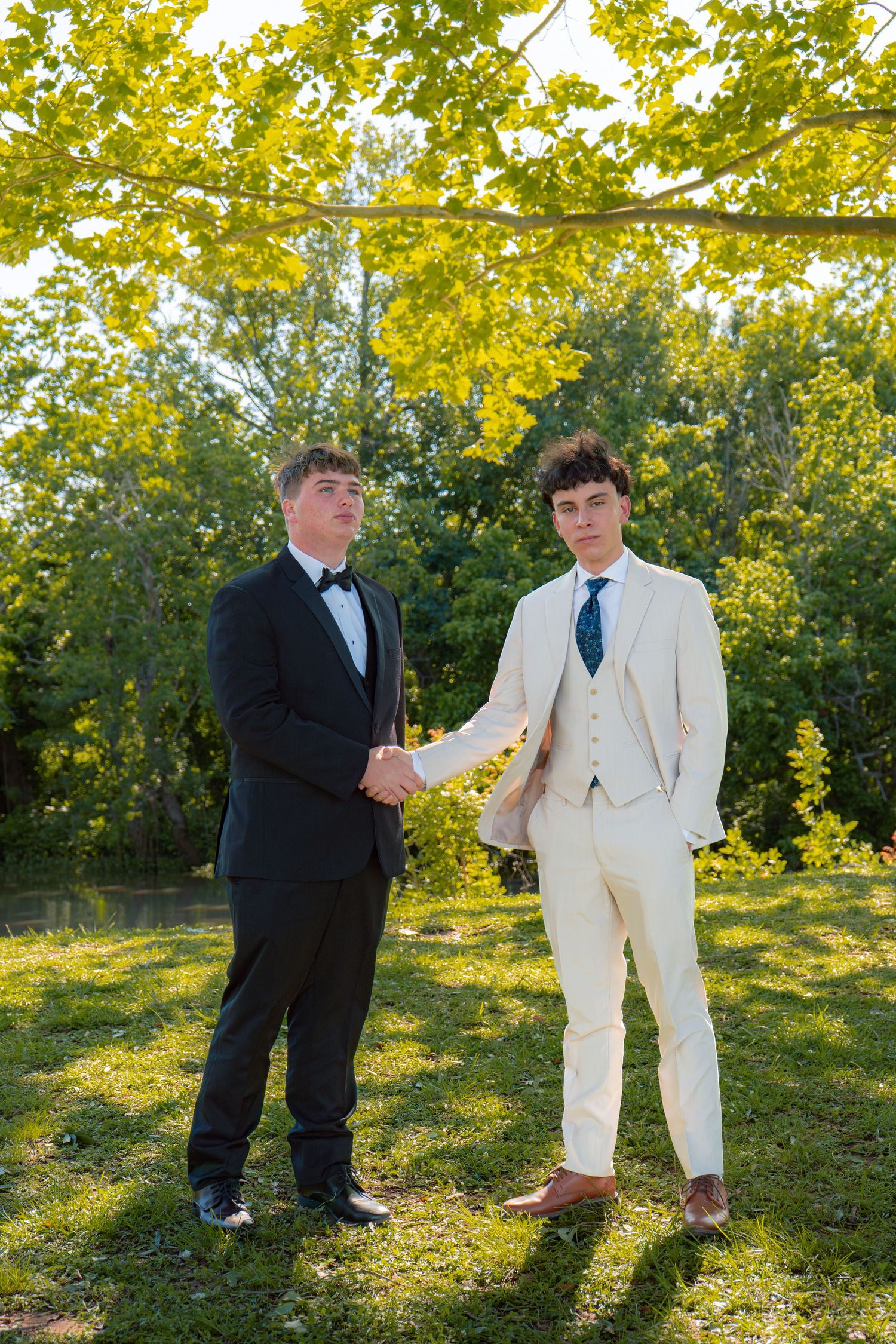 Two young men in formal suits shake hands outdoors under bright yellow-green tree branches at prom.