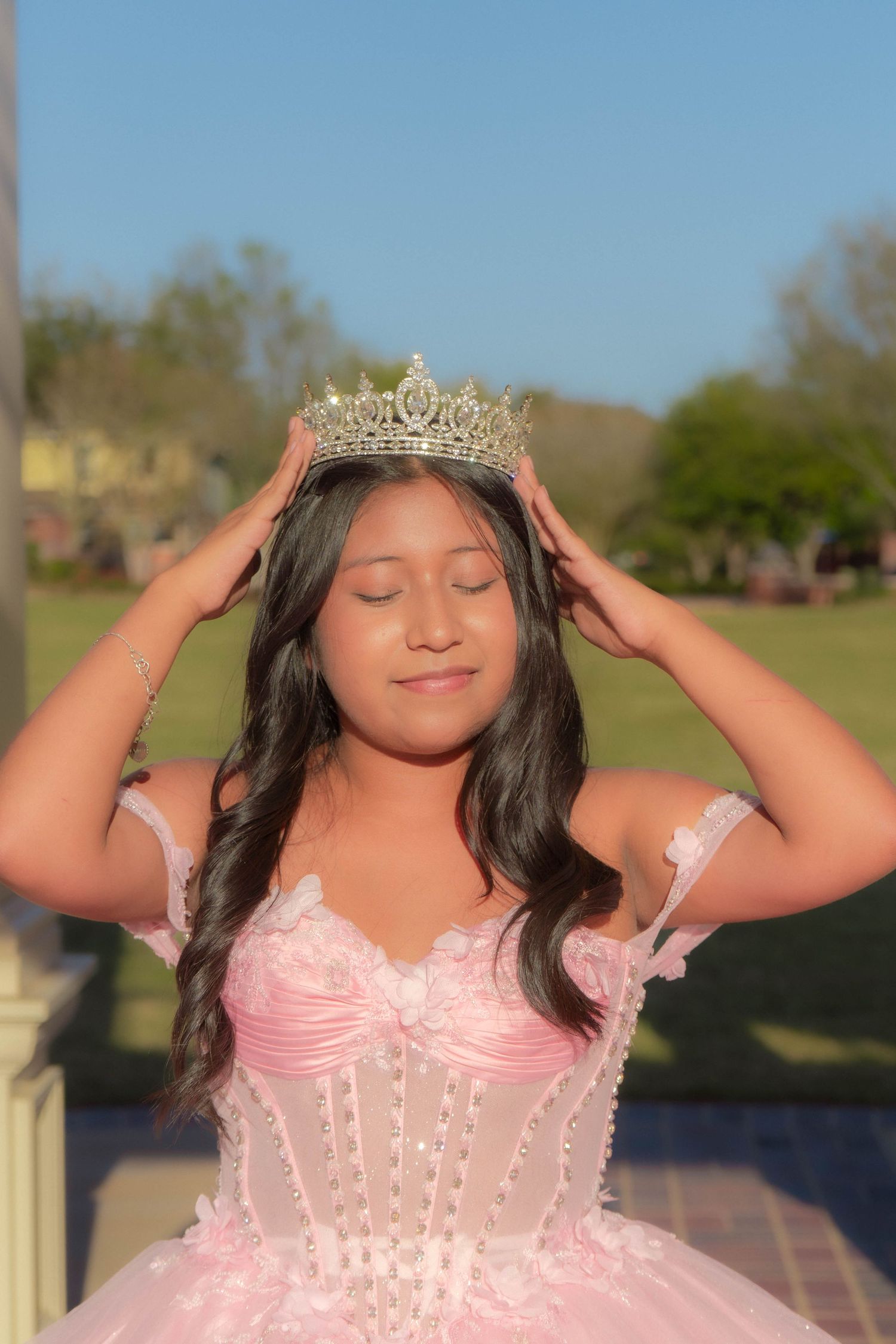 Young Latina girl in pink quinceañera gown placing silver tiara crown on her head outdoors.