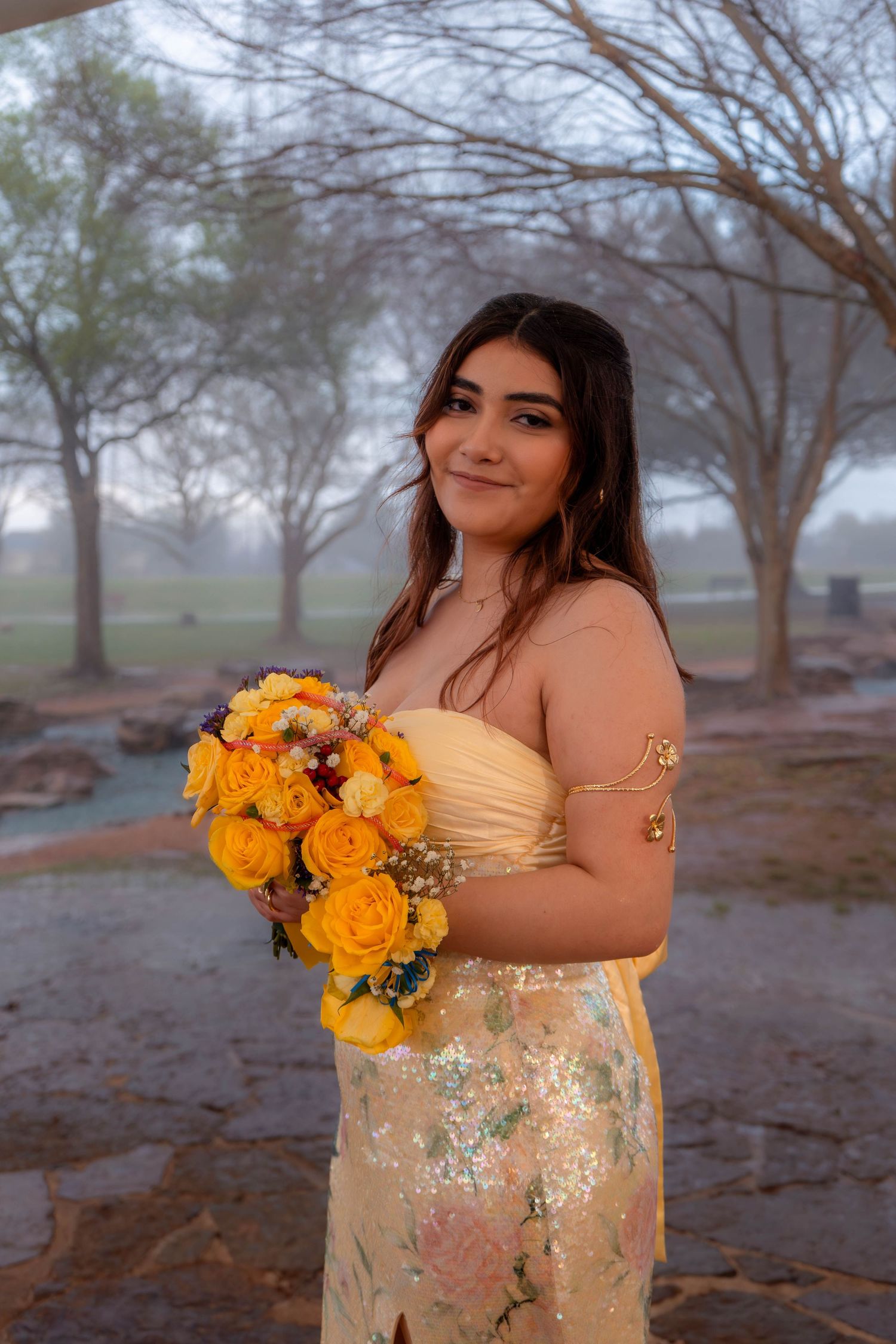 Young woman in gold sequin strapless prom gown smiling while holding a bouquet of yellow roses in a misty park setting.