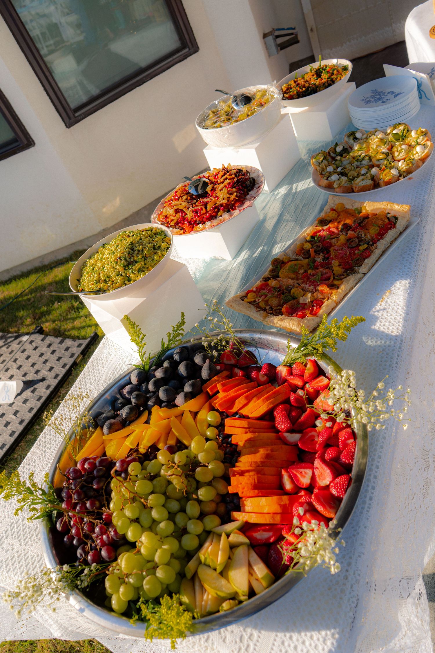 Outdoor baby shower buffet table with colorful fruit platter, grain salads, vegetable tart, and assorted dishes on white linen.