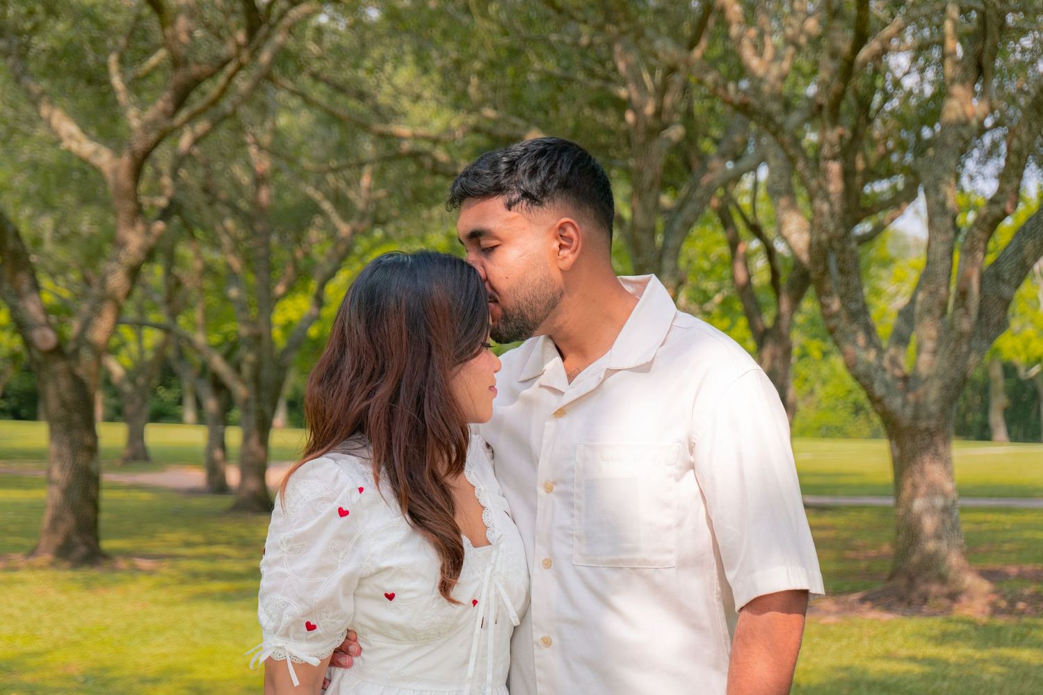 Couple sharing a romantic kiss in a lush green park, both wearing white outfits on a sunny day.