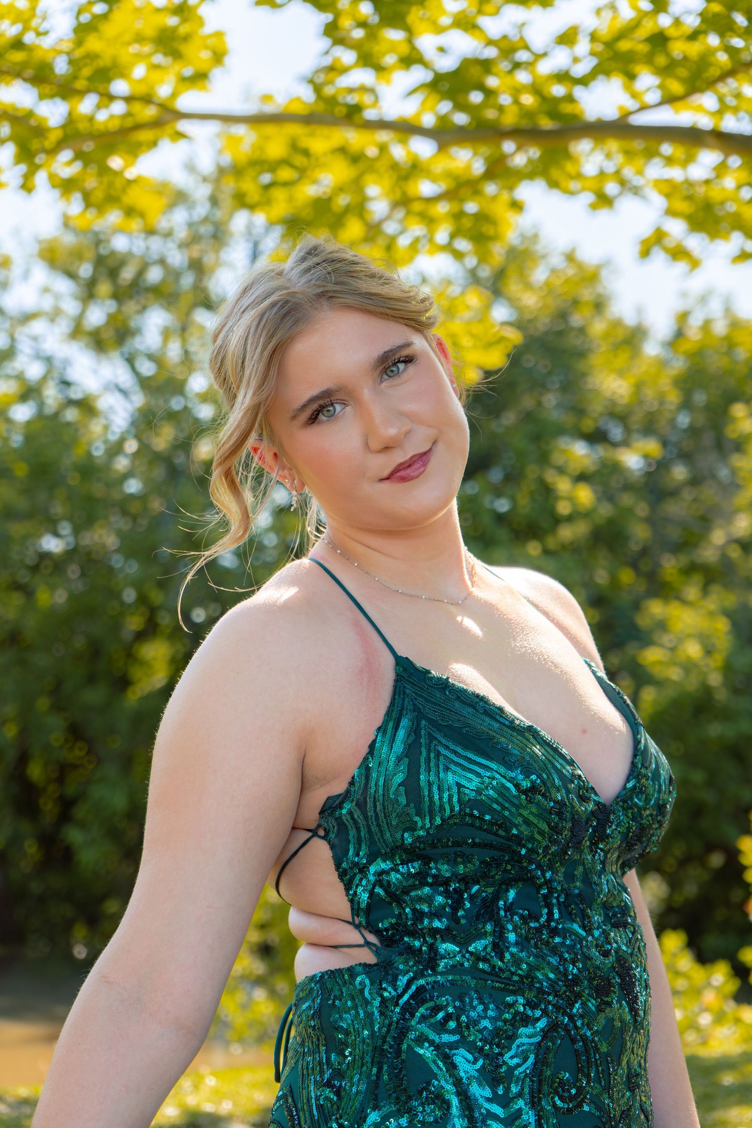 Young blonde woman in a deep green sequined formal gown poses outdoors under bright yellow-green tree foliage.