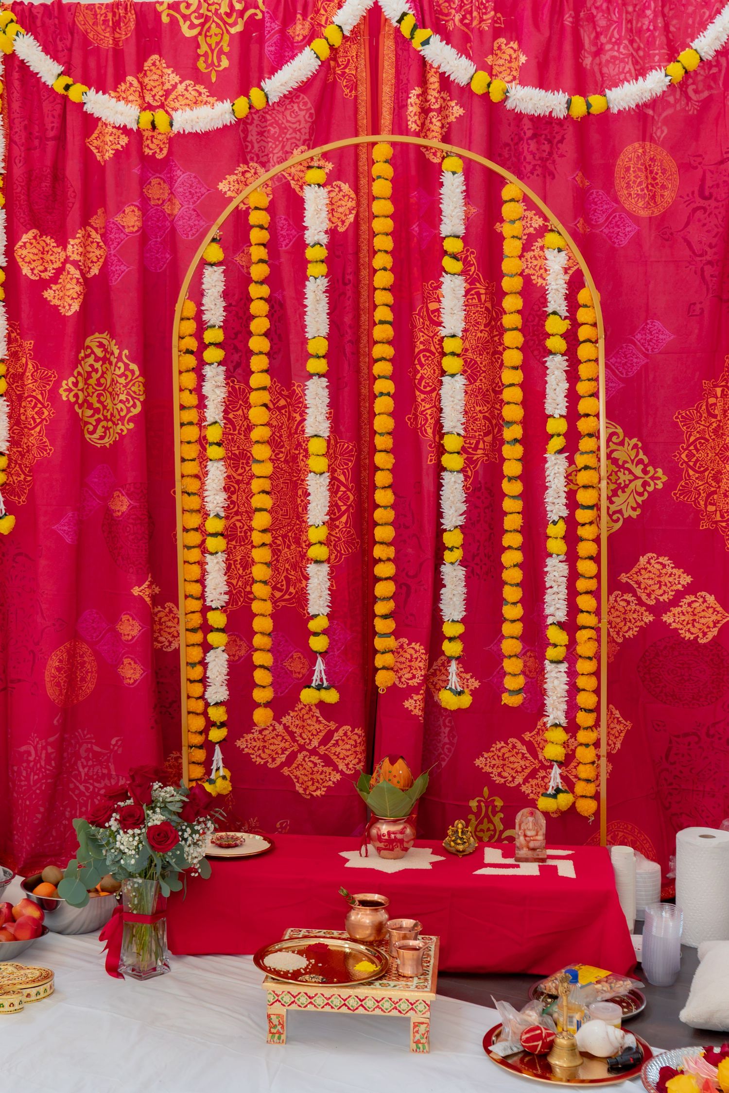 Traditional Indian puja setup with marigold garlands, red backdrop, and ceremonial offerings on decorated altar.
