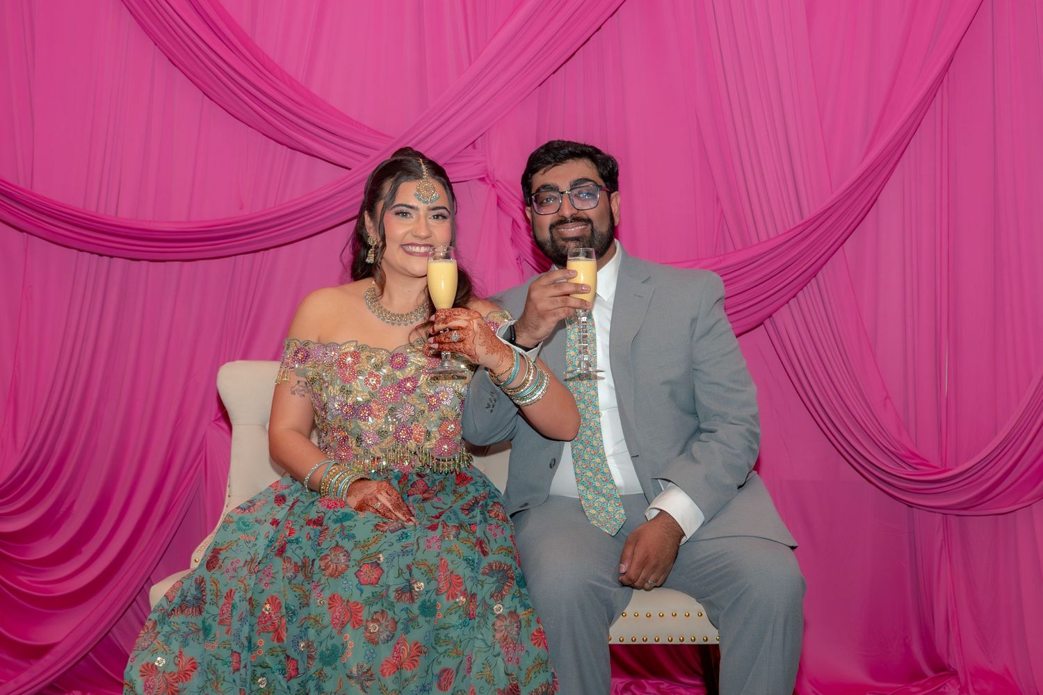 Couple in formal attire toasting with champagne glasses against a pink draped backdrop at a celebration.