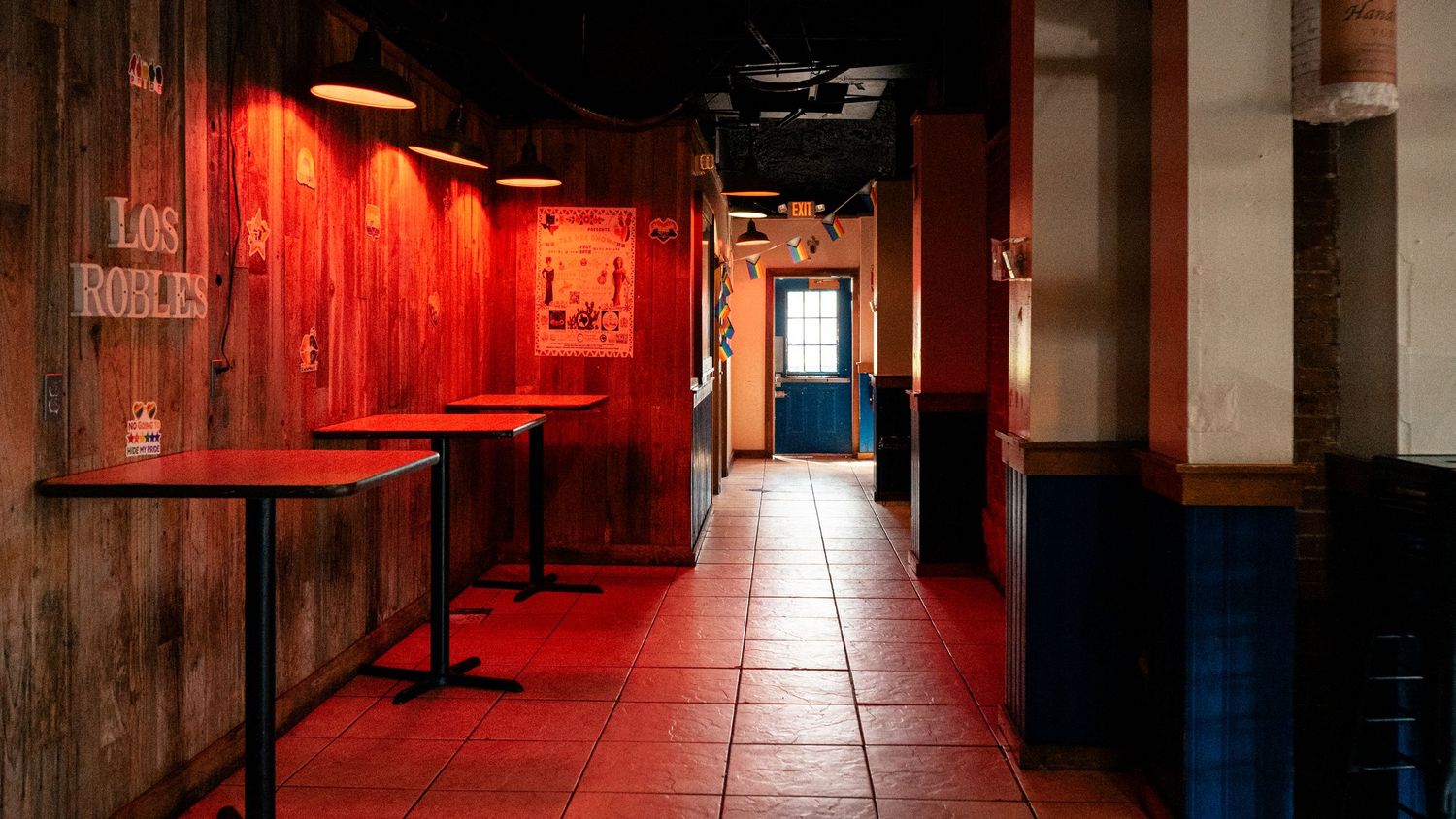 Interior of Los Robles bar with red lighting, wooden walls, high tables, and tiled floor.