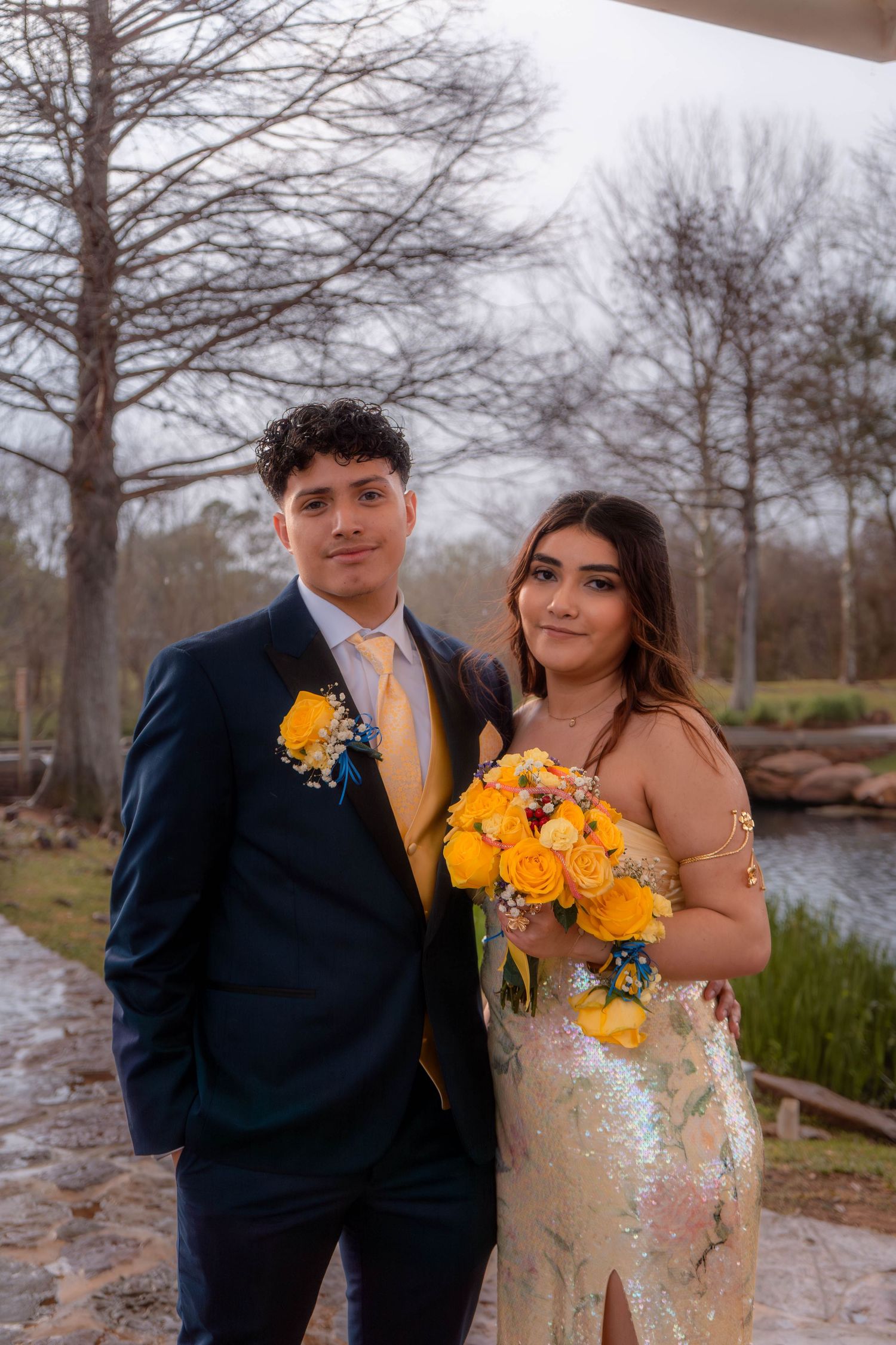 Prom couple posing together near a pond, young man in navy suit with yellow tie and girl in gold sequin dress with yellow roses.