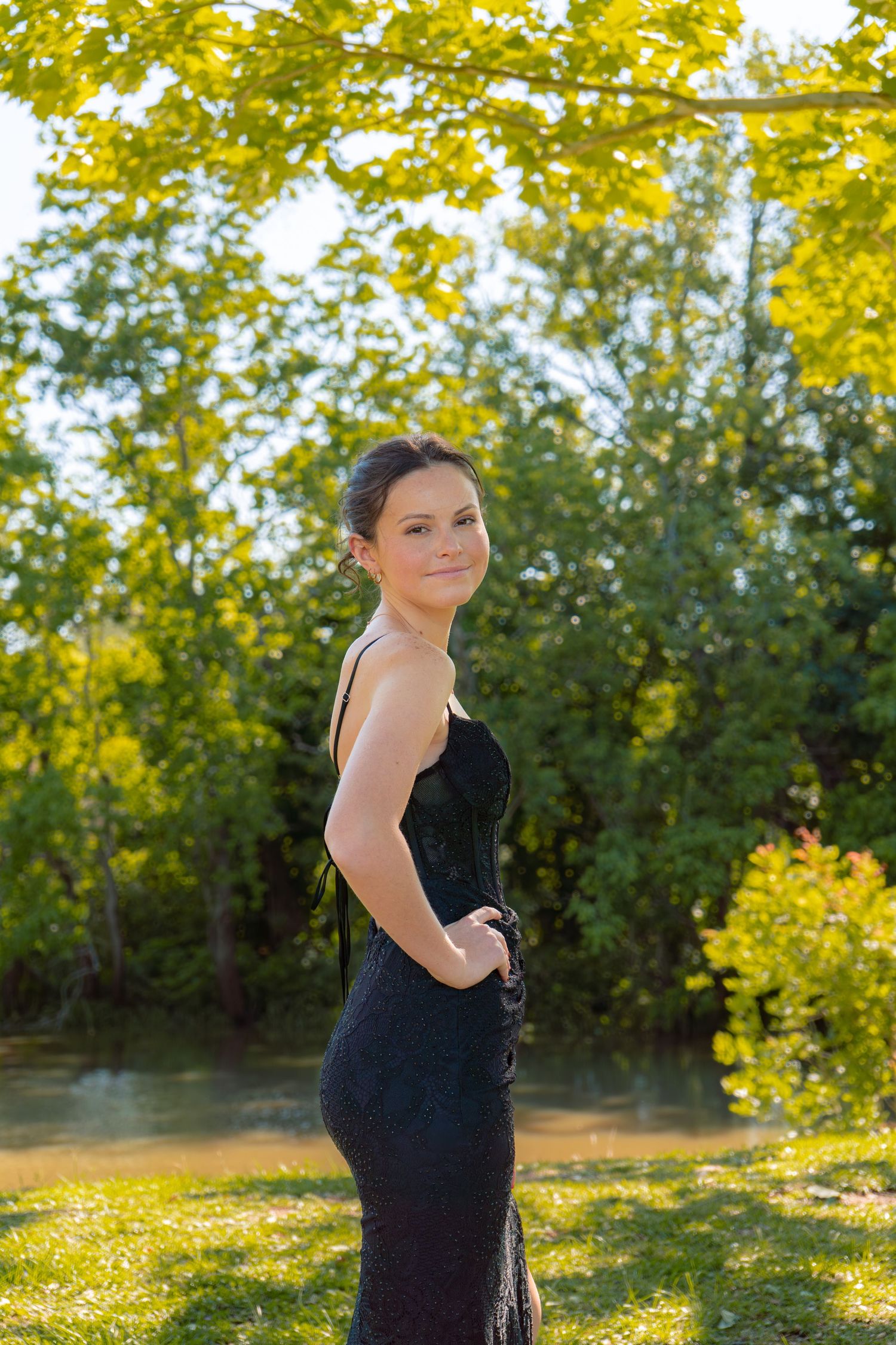 Young woman in a black sequin prom dress poses outdoors near a pond surrounded by green trees.
