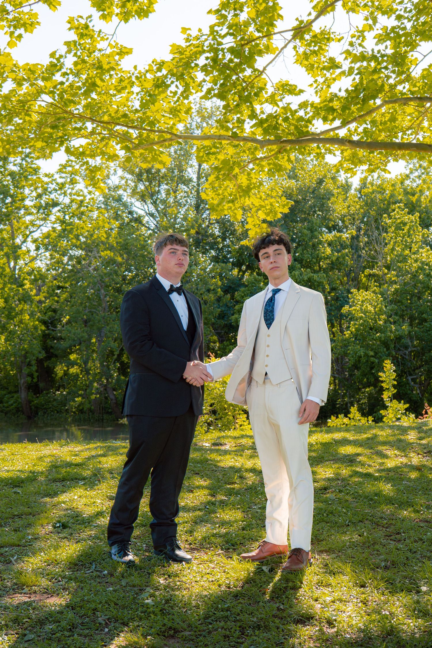 Two young men in formal suits shake hands outdoors under bright yellow-green tree branches on a sunny day.