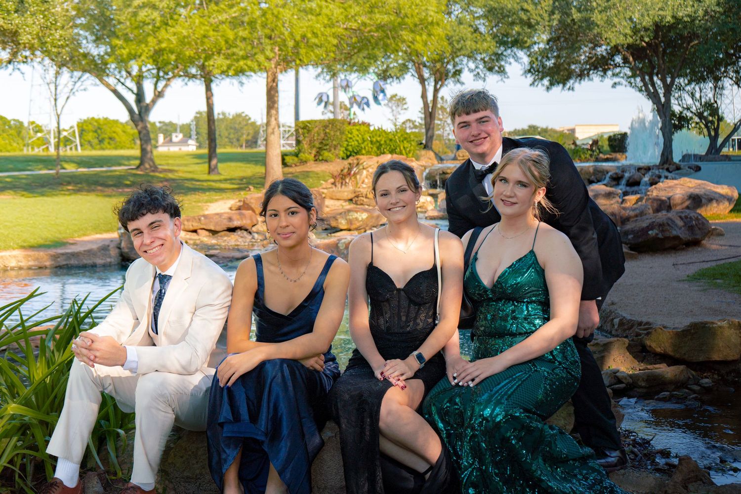 Five teenagers dressed in formal prom attire sit together outdoors near a decorative garden fountain.
