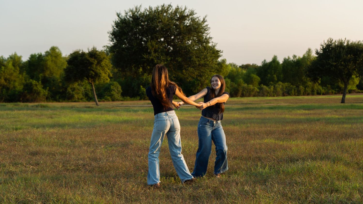 Two young women in jeans dancing together in a green field at sunset with trees in the background.