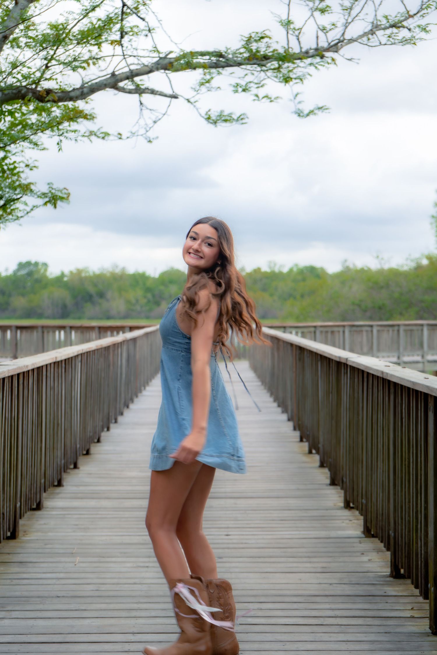 Young woman in blue dress and brown boots smiling on wooden boardwalk near water.
