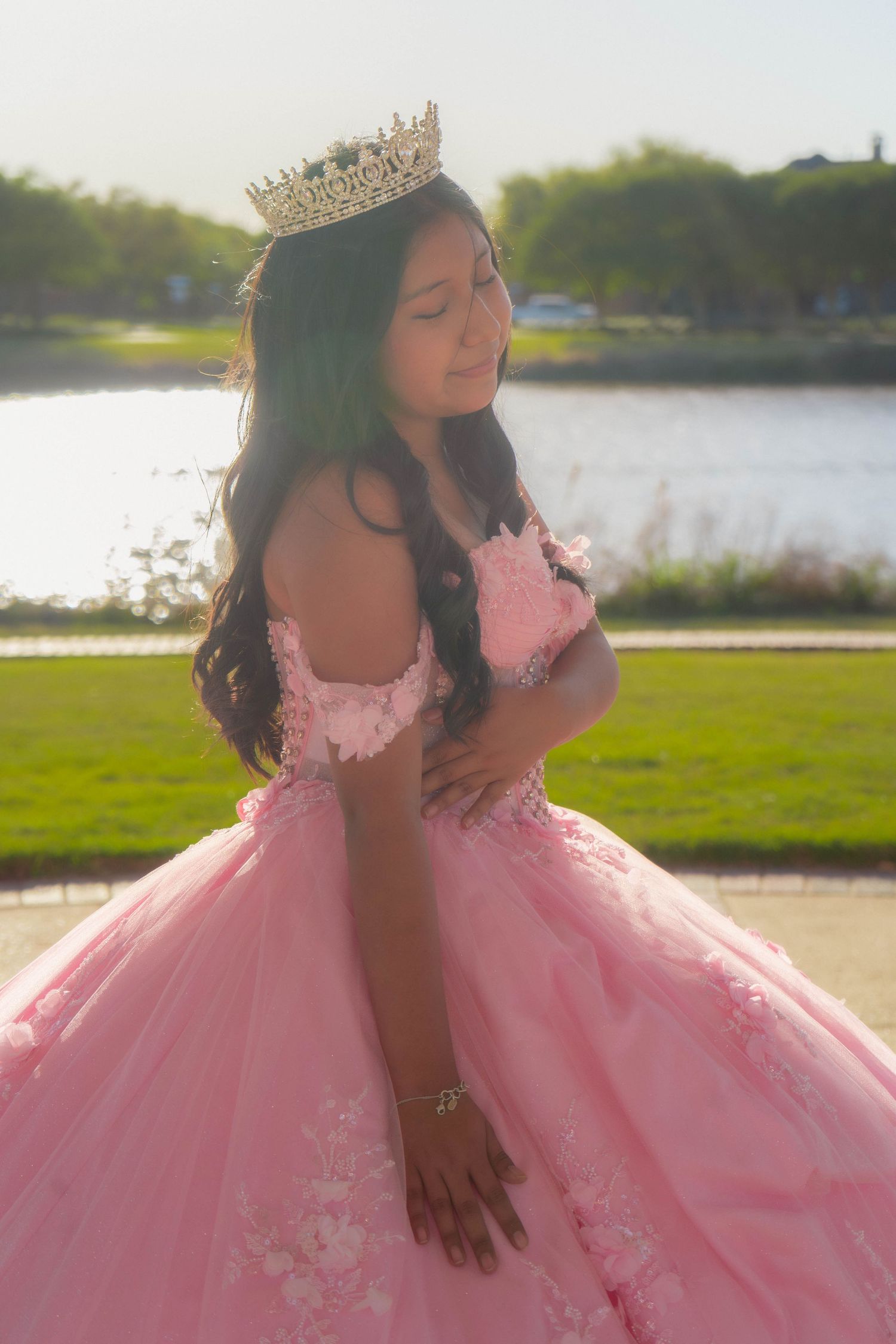 Young Latina girl in pink quinceañera ball gown and gold tiara poses gracefully outdoors near a lake.