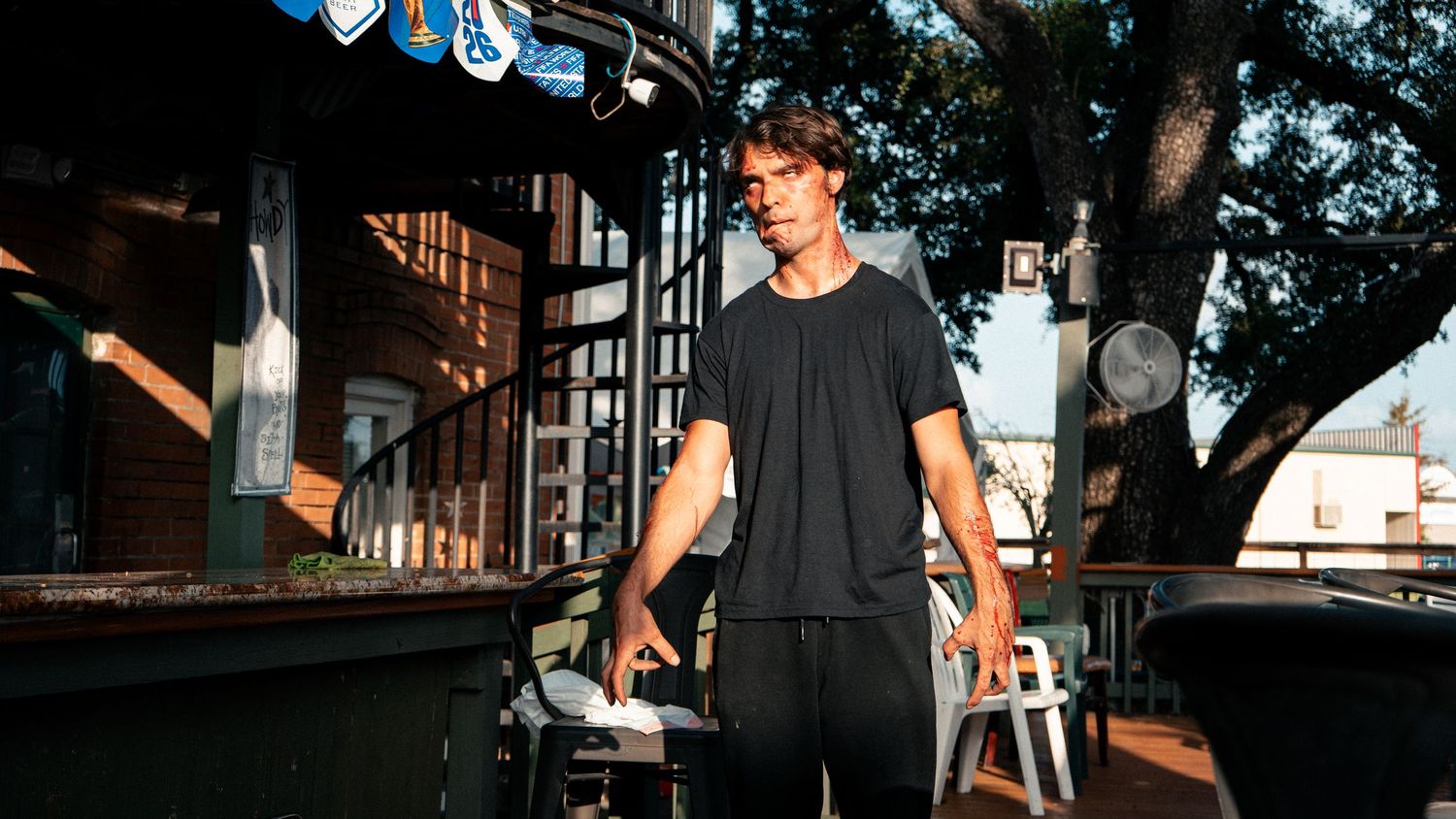 A man stands outdoors on a sunny patio near a bar with trees in background.