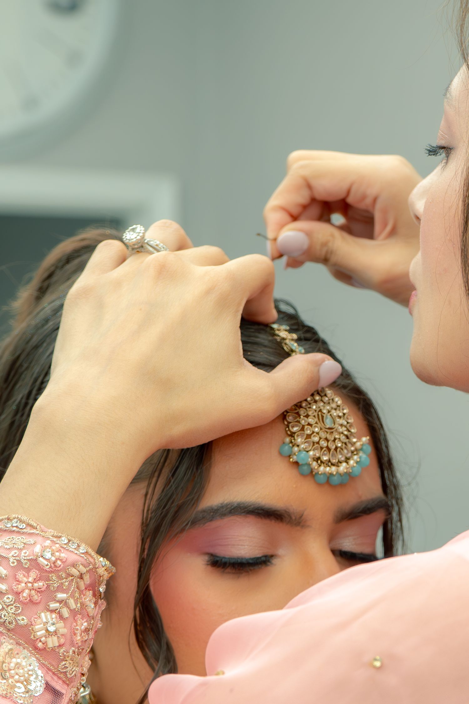 Makeup artist applying a traditional maang tikka headpiece on a bride wearing pink Indian bridal attire.